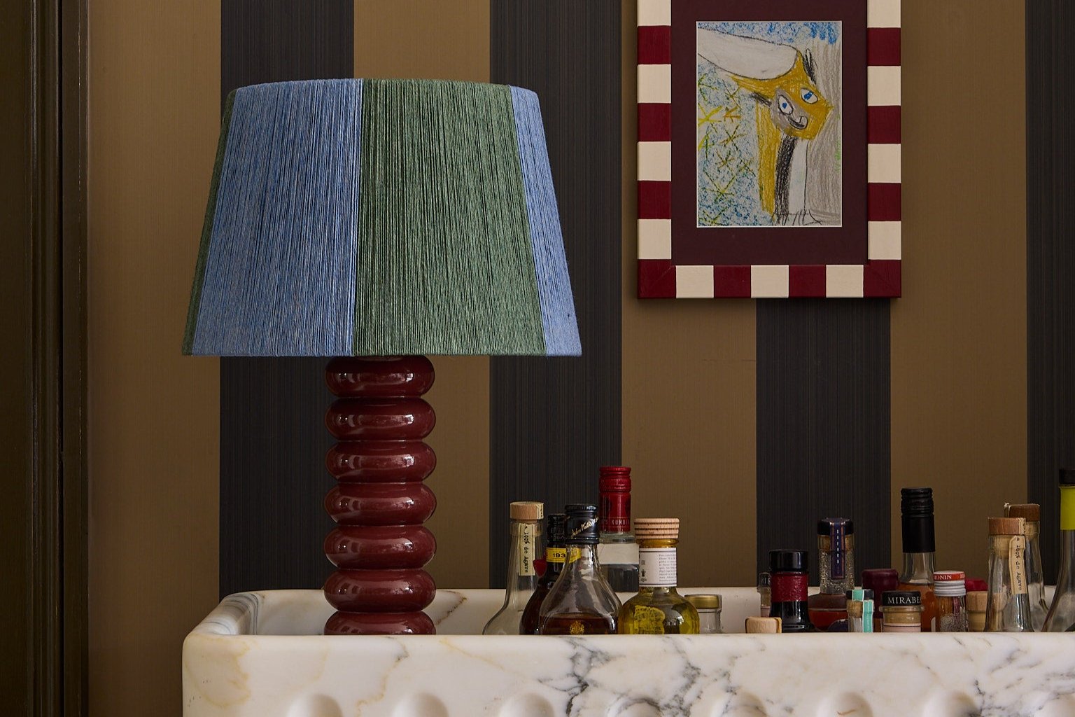 A blue and green stripey string lampshade on a red lamp on a counter top in front of a striped wall