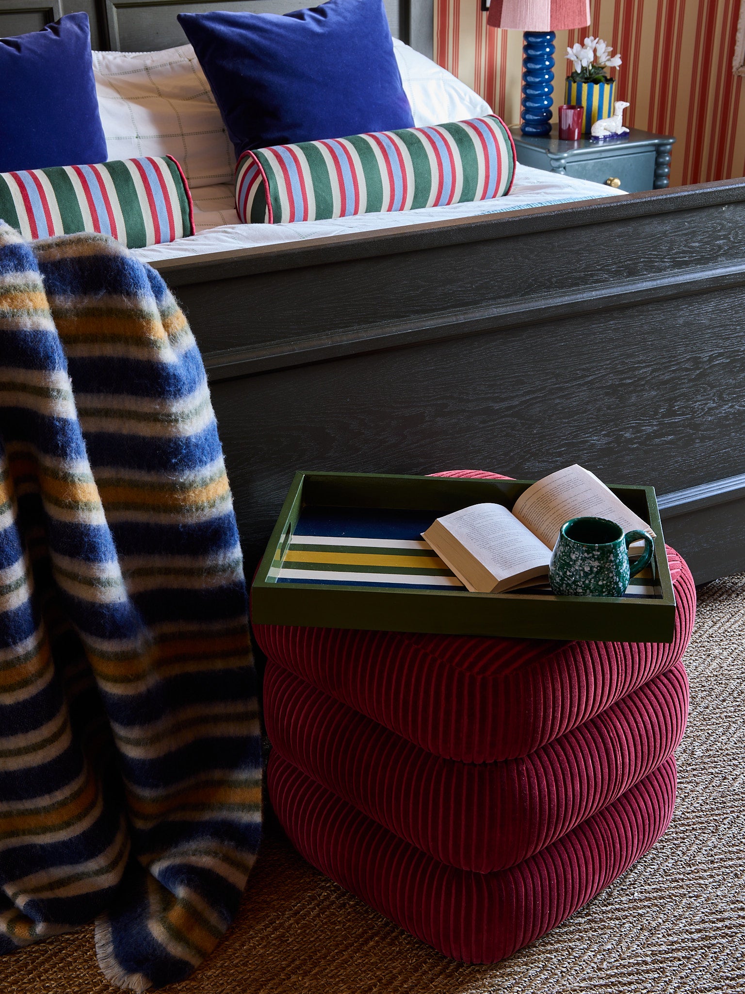 A very good tray rests on the classic stool with an open book, next to a striped throw draped over the bed