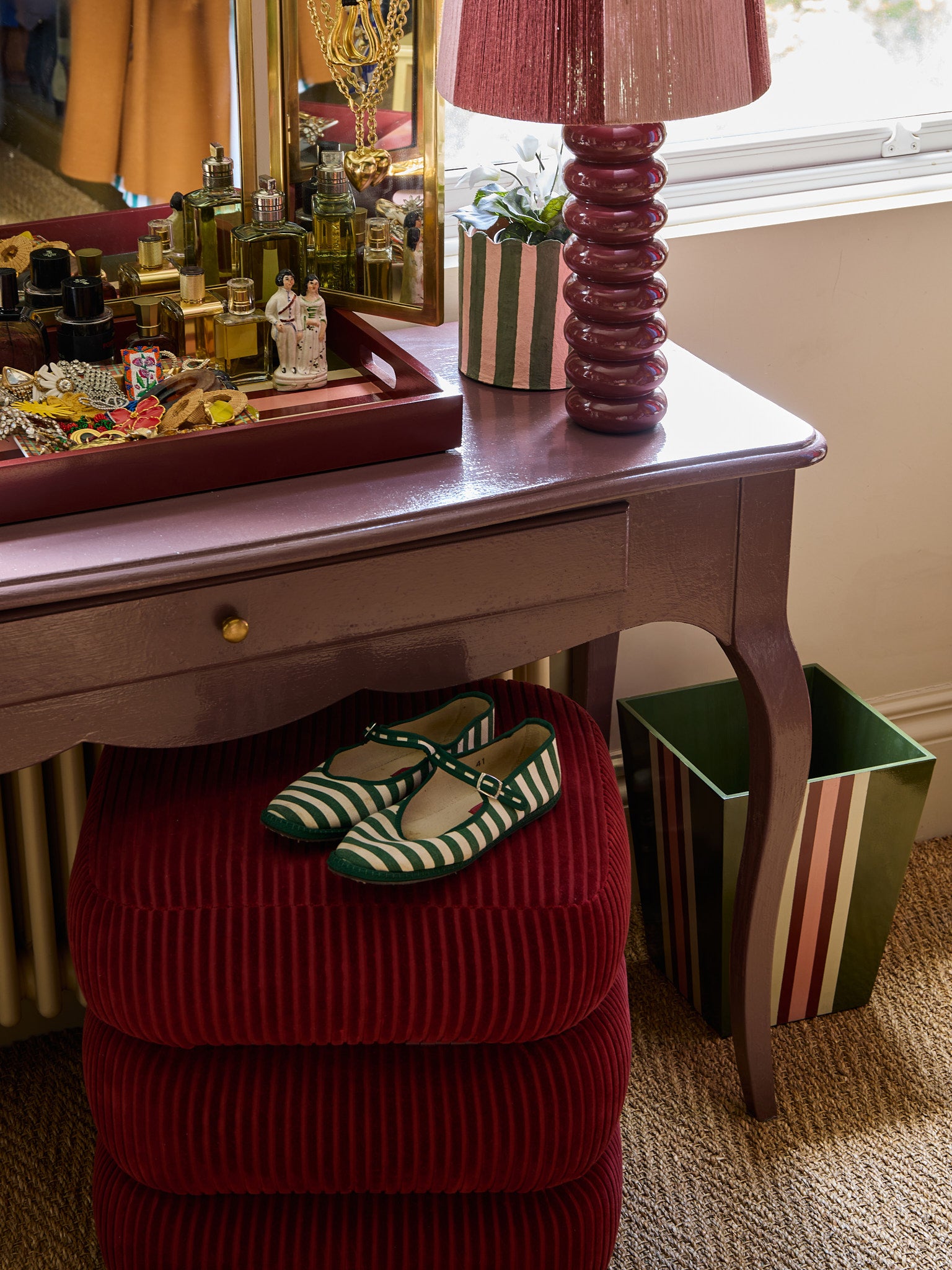 A pair of green and white shoes sit on the classic stool in red, tucked under a dressing table. The dressing table has a lamp and a tray on top.