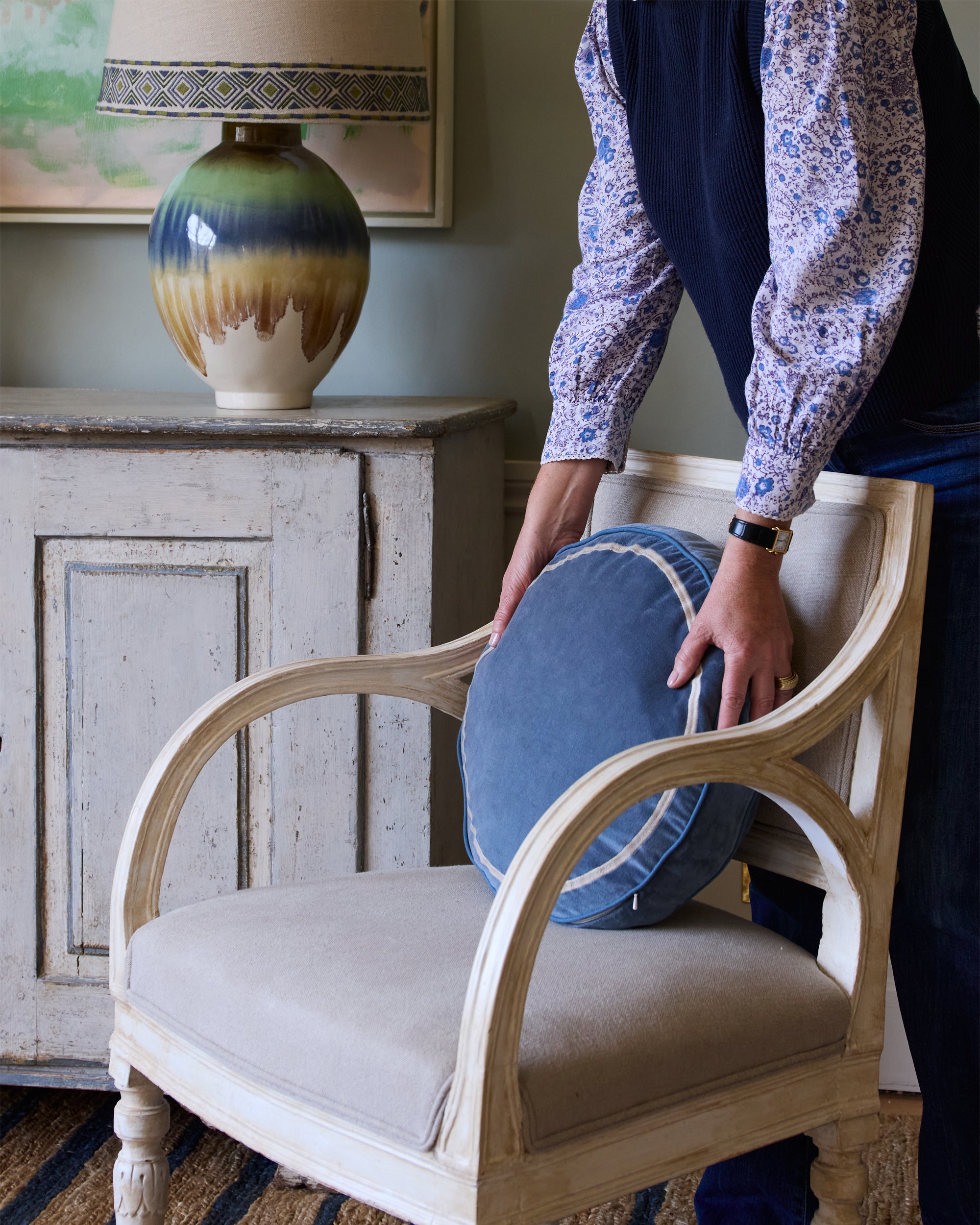 Person adjusting a blue cushion on a beige armchair in a room with a lamp and cabinet.