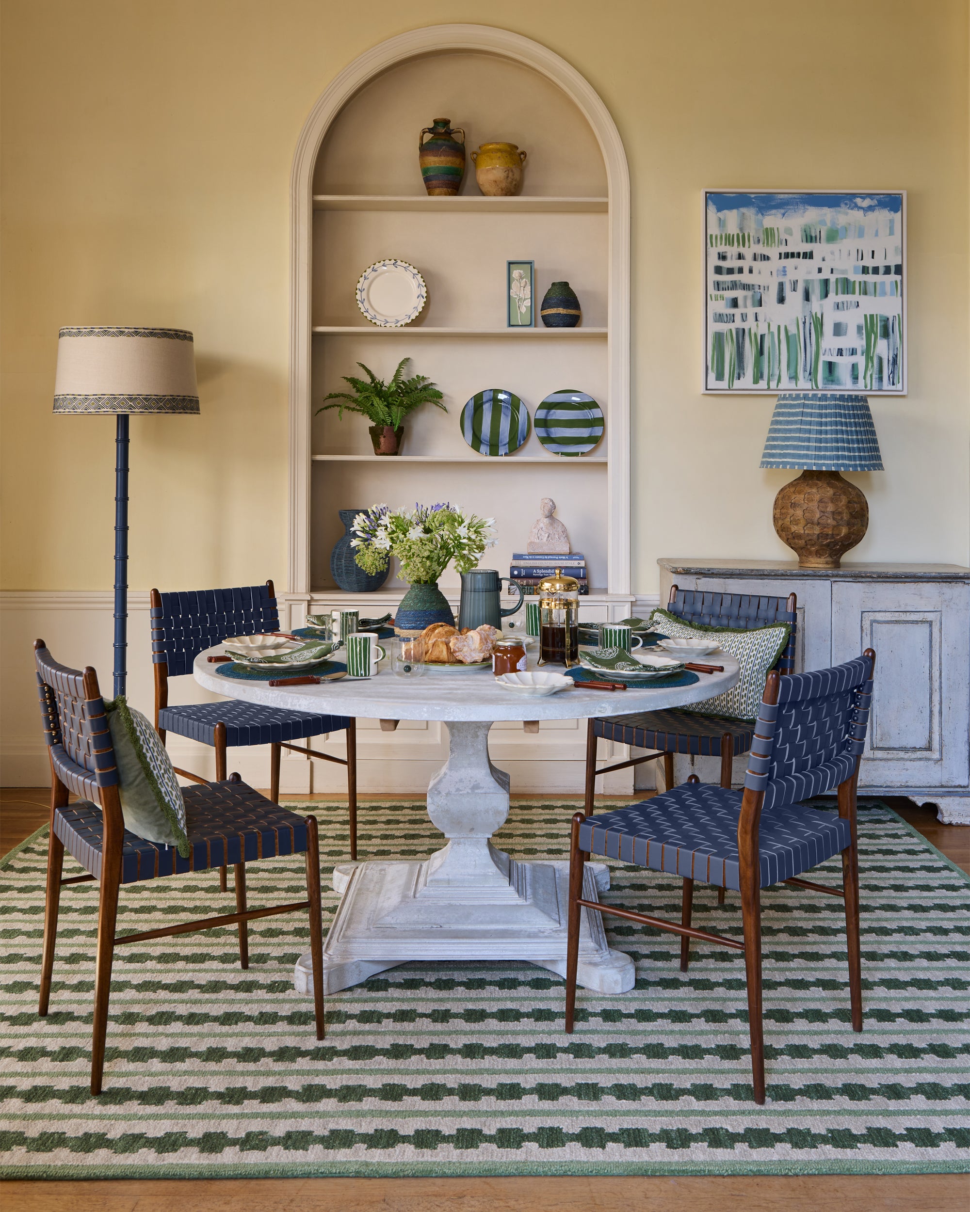 Dining room with a round table and blue chairs, featuring a shelf with decorative items and a rug.
