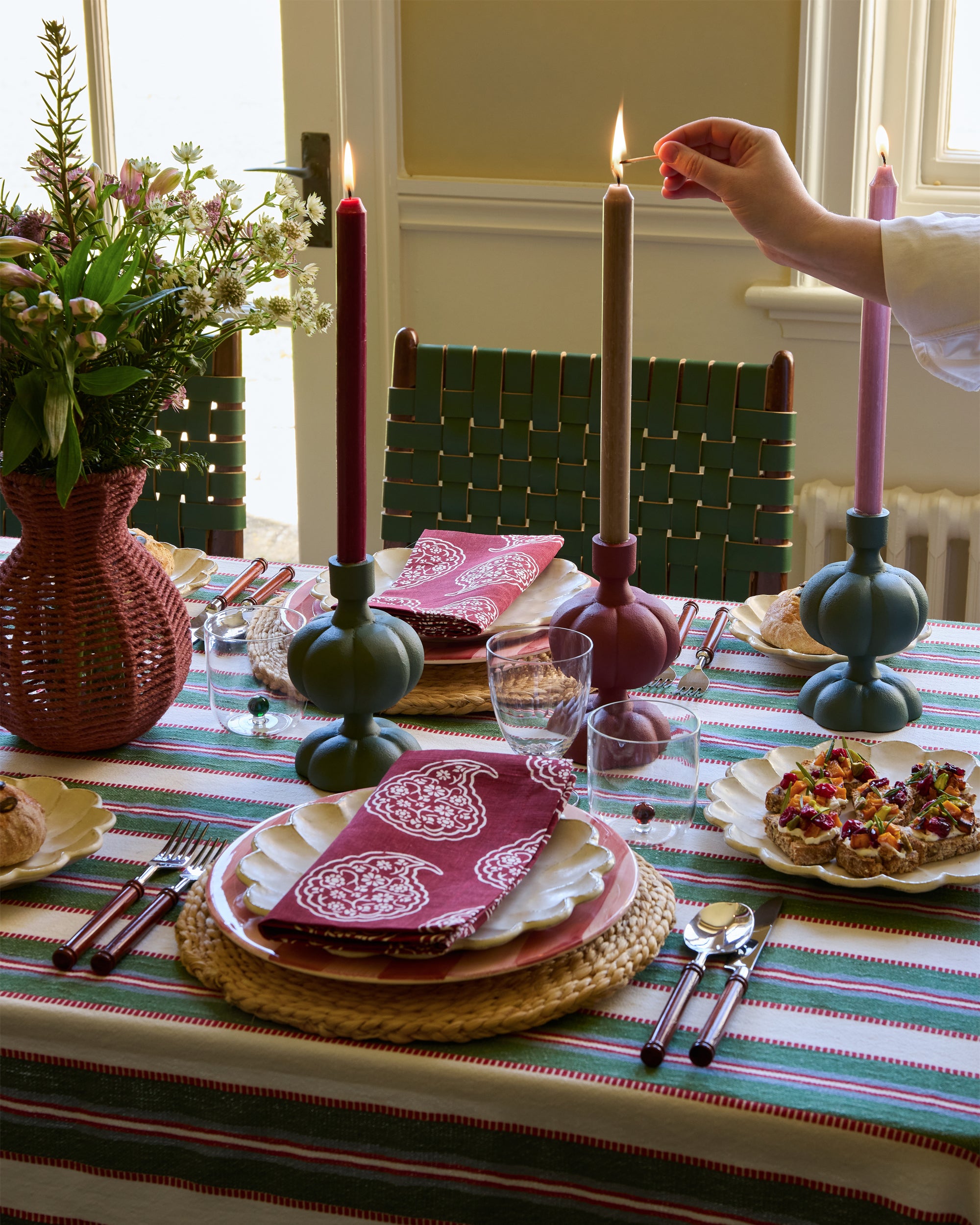 Person lighting a candle on a decorated dining table with colorful tablecloth and floral arrangement.