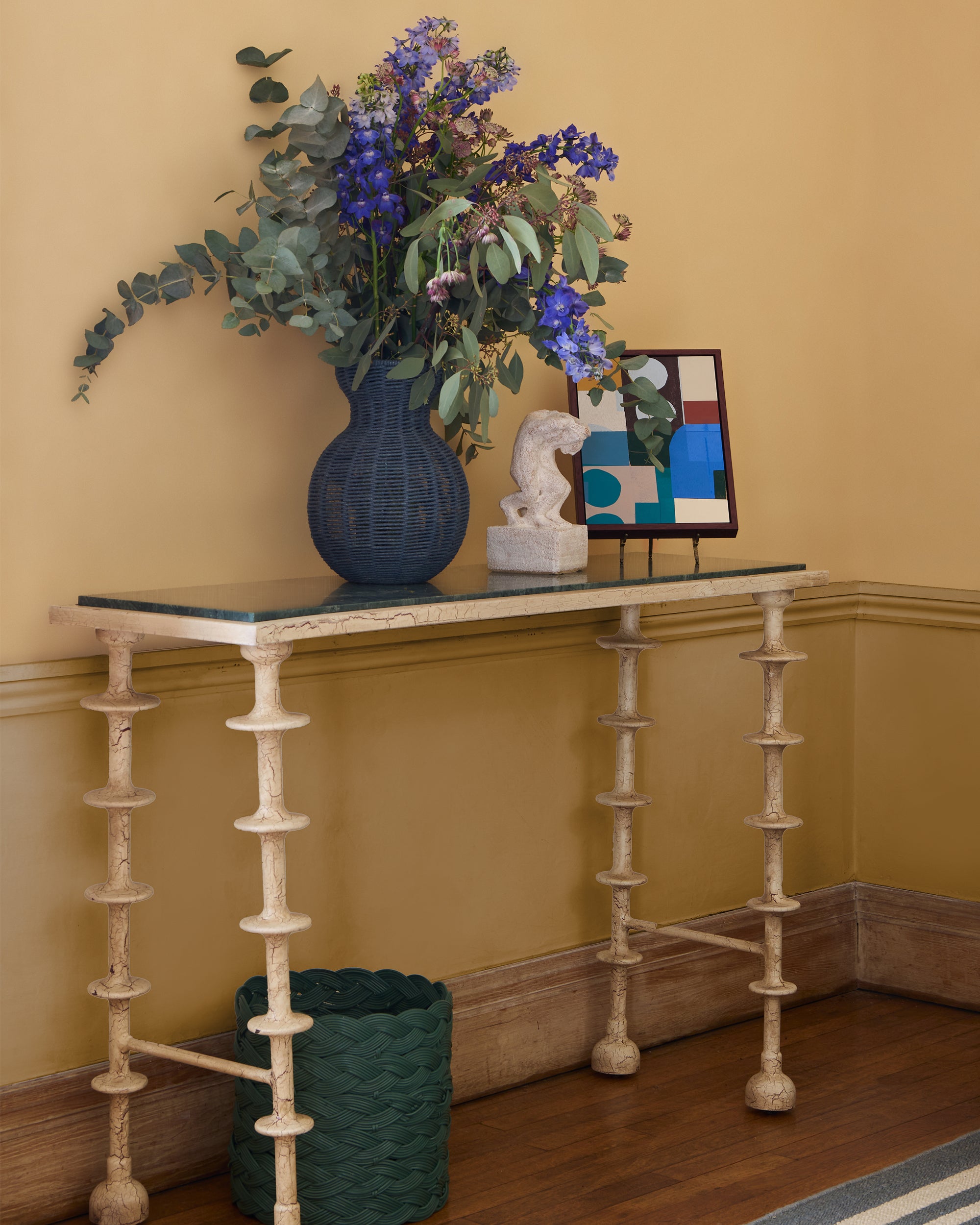 Decorative console table with a vase of flowers, sculpture, and abstract painting against a beige wall.