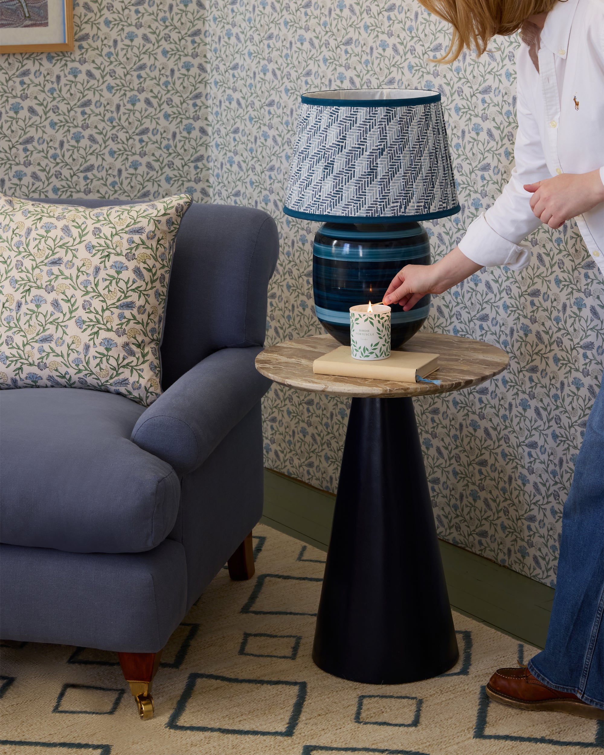 Person lighting a candle on a small round table next to a blue armchair with floral-patterned cushions.