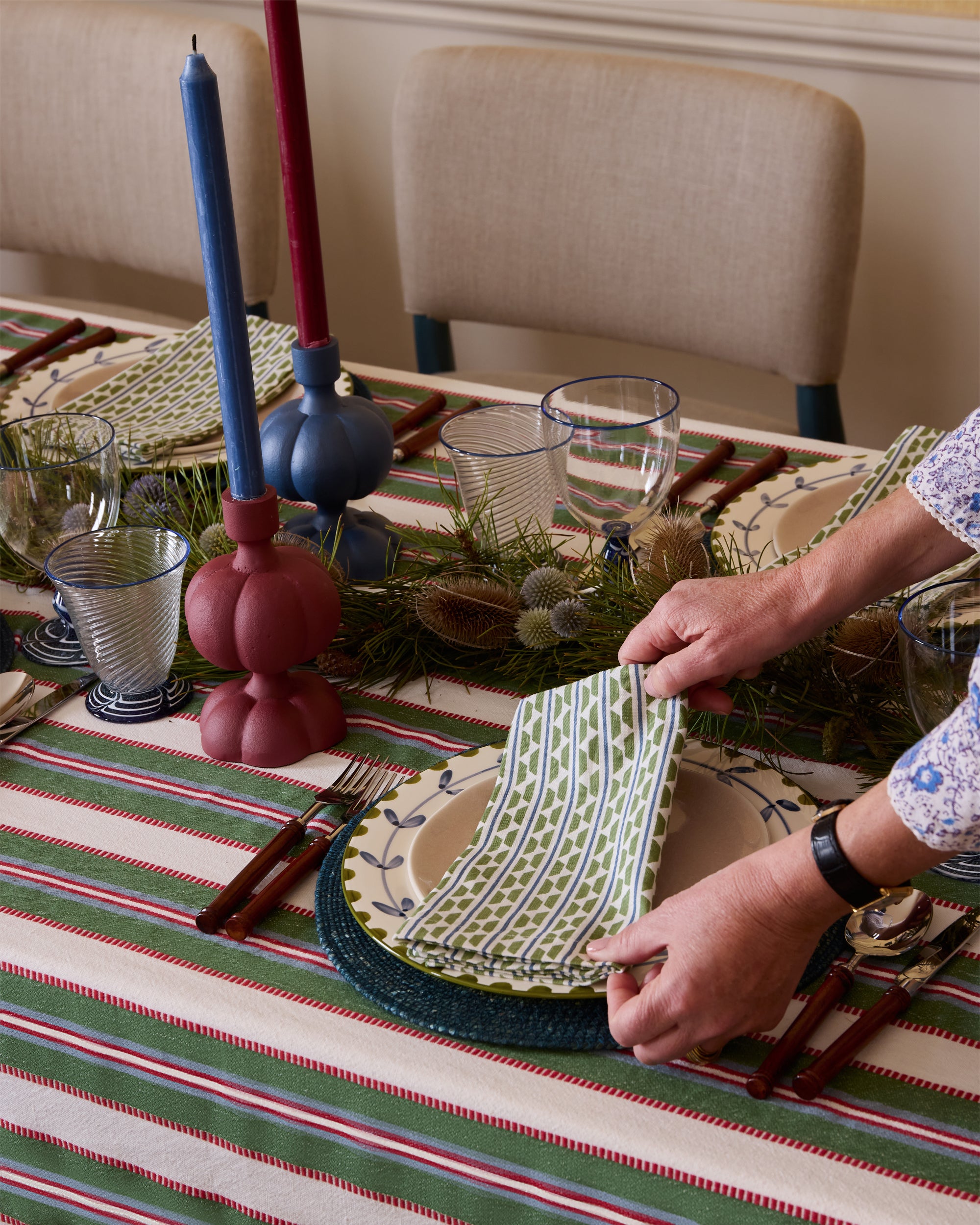Person setting a table with decorative plates and candles on a striped tablecloth.