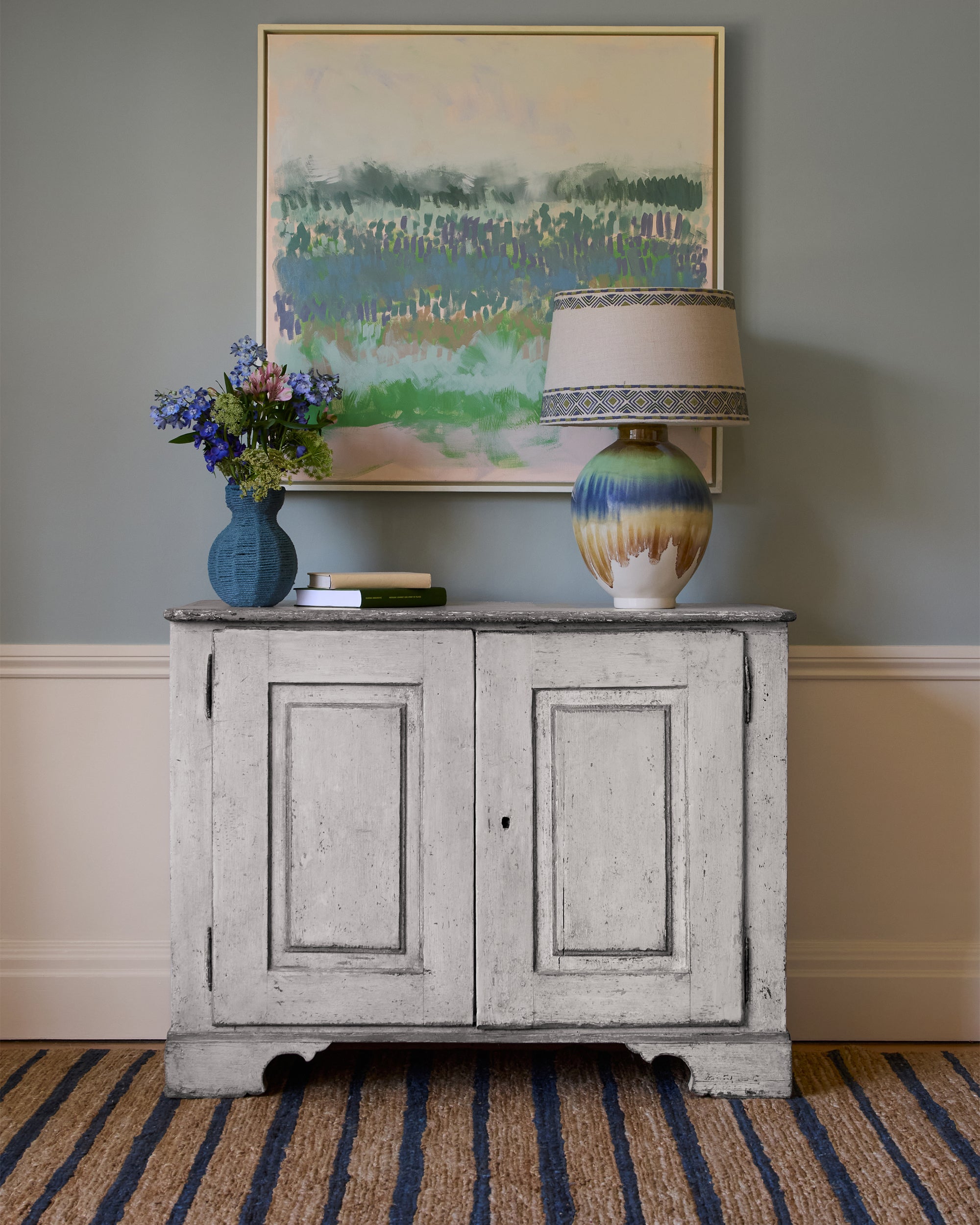 Vintage-style sideboard with a lamp, vase, and flowers against a wall with a framed landscape painting.