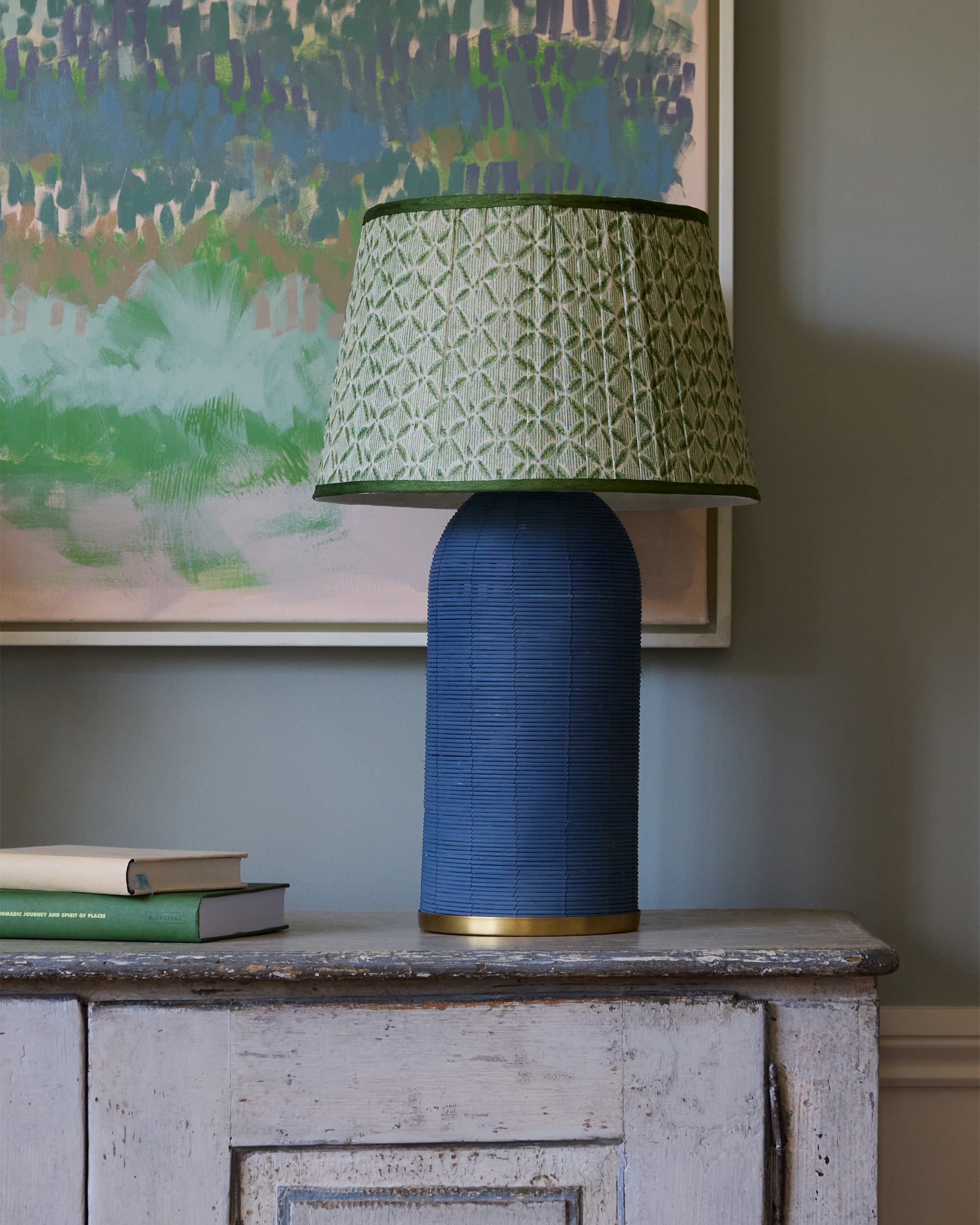 Blue textured table lamp with a green patterned shade on a wooden surface, with a colorful abstract painting in the background.