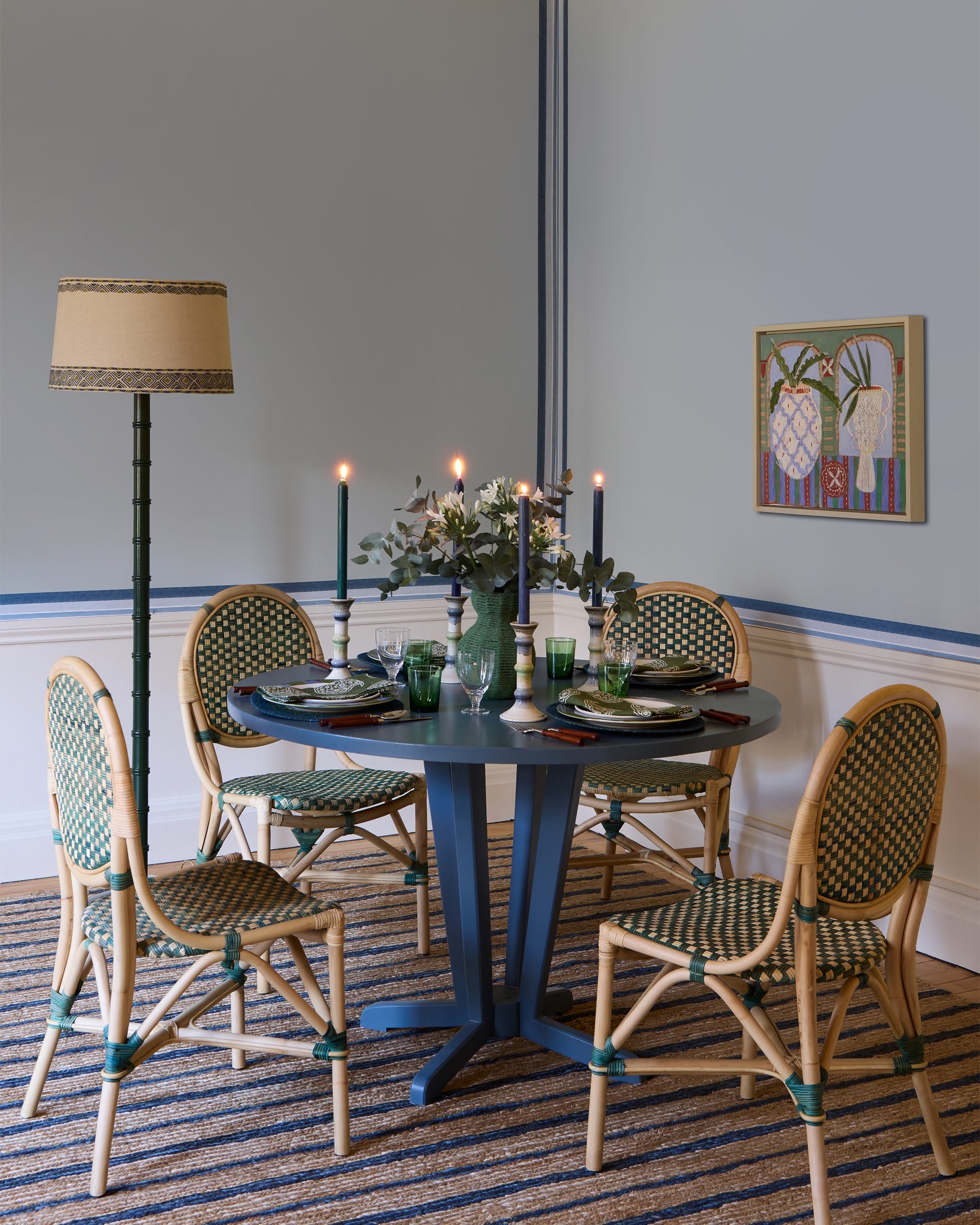 Dining room with a blue table and wicker chairs, decorated with candles and a vase.