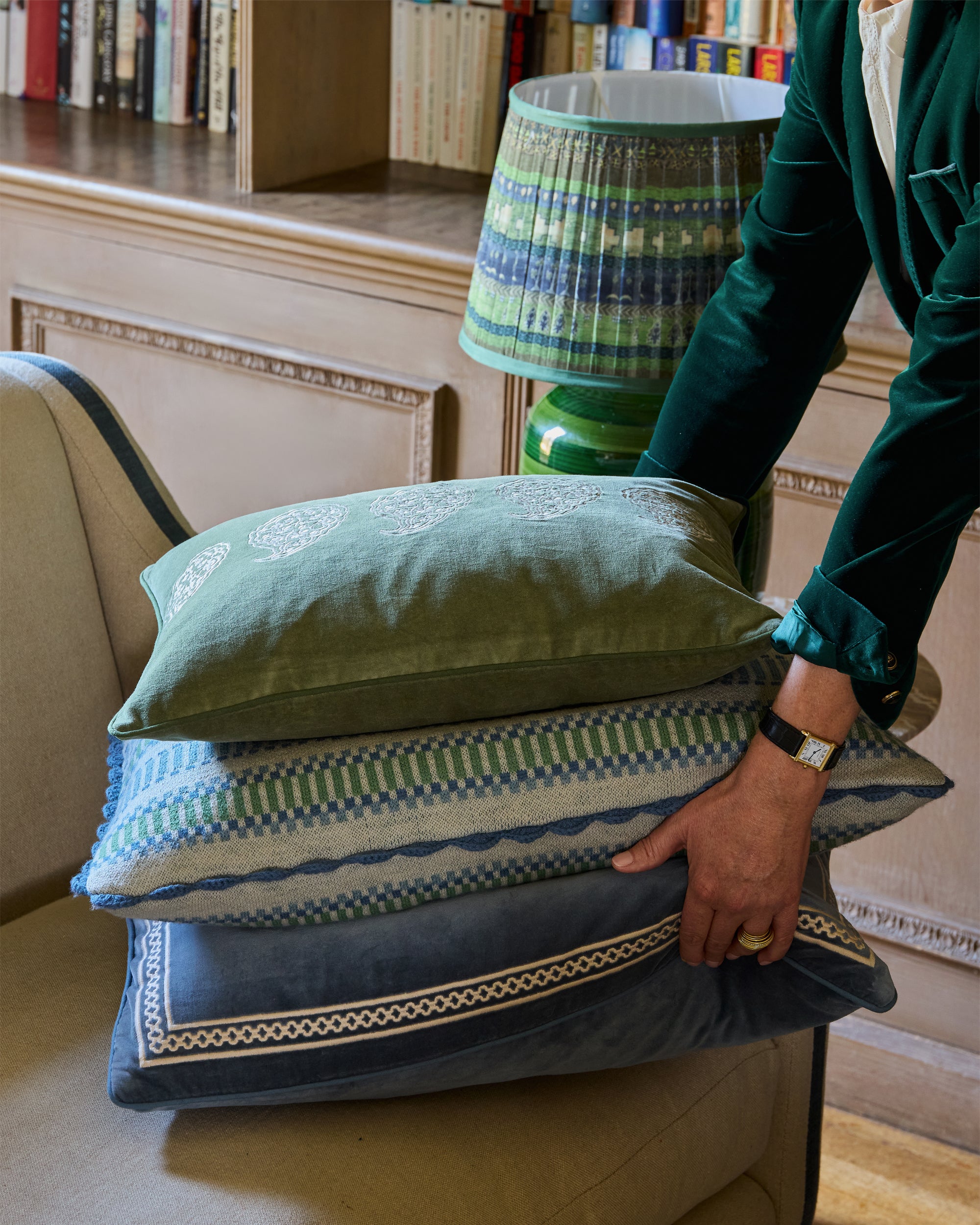 Person arranging decorative pillows on a chair with a lamp in the background