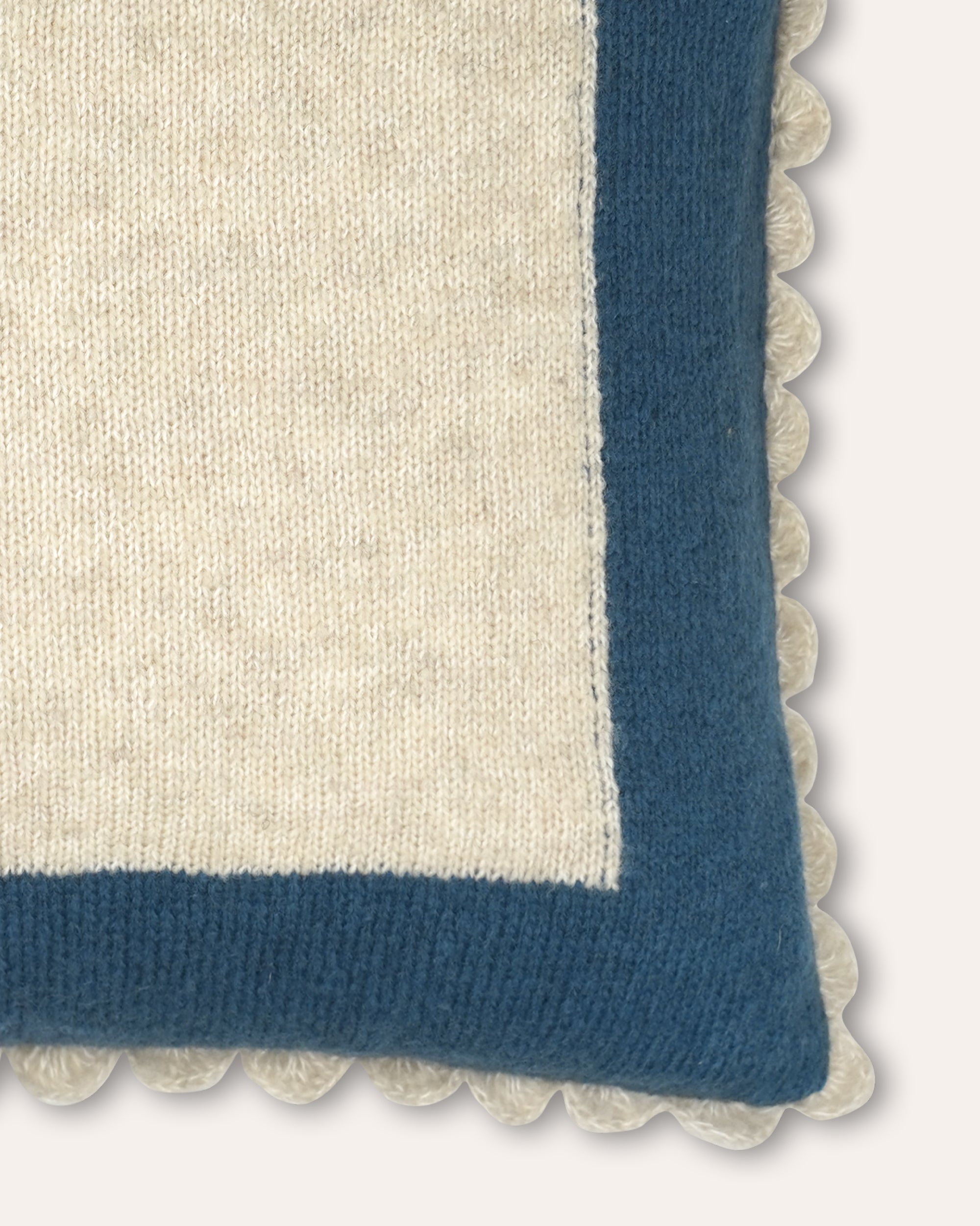Close-up of a knitted throw pillow corner showing a cream center, a blue border, and a cream pom-pom trim on a light neutral background.