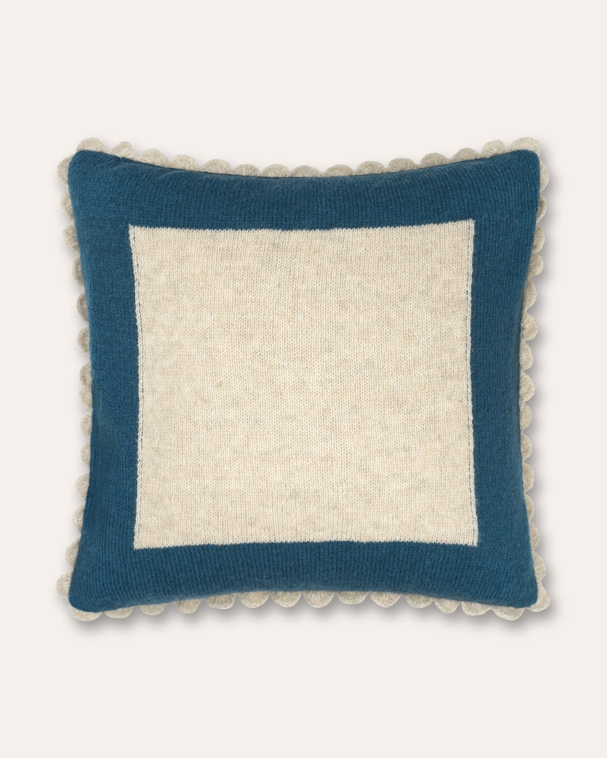 A square knit cushion with a deep teal blue border and a cream center panel, finished with a cream pom-pom trim around the edge, shown against a light beige background.