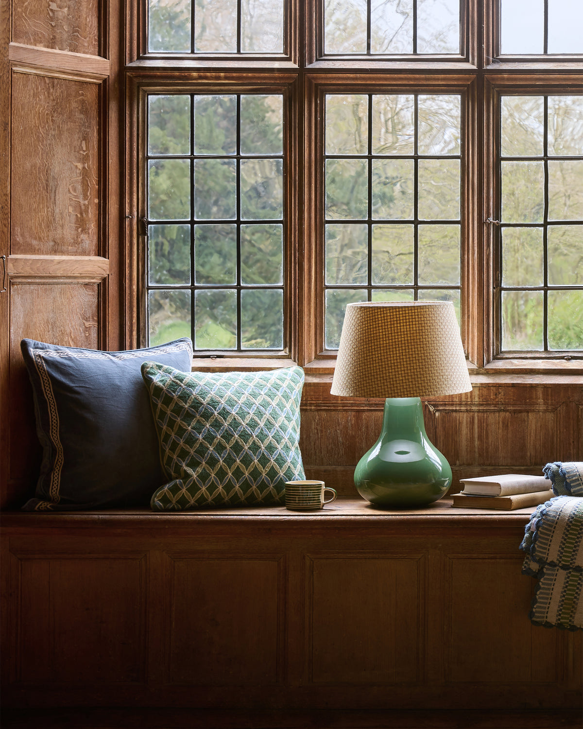 Nook with cushions, a lamp, and books by large windows in a room with wooden walls.