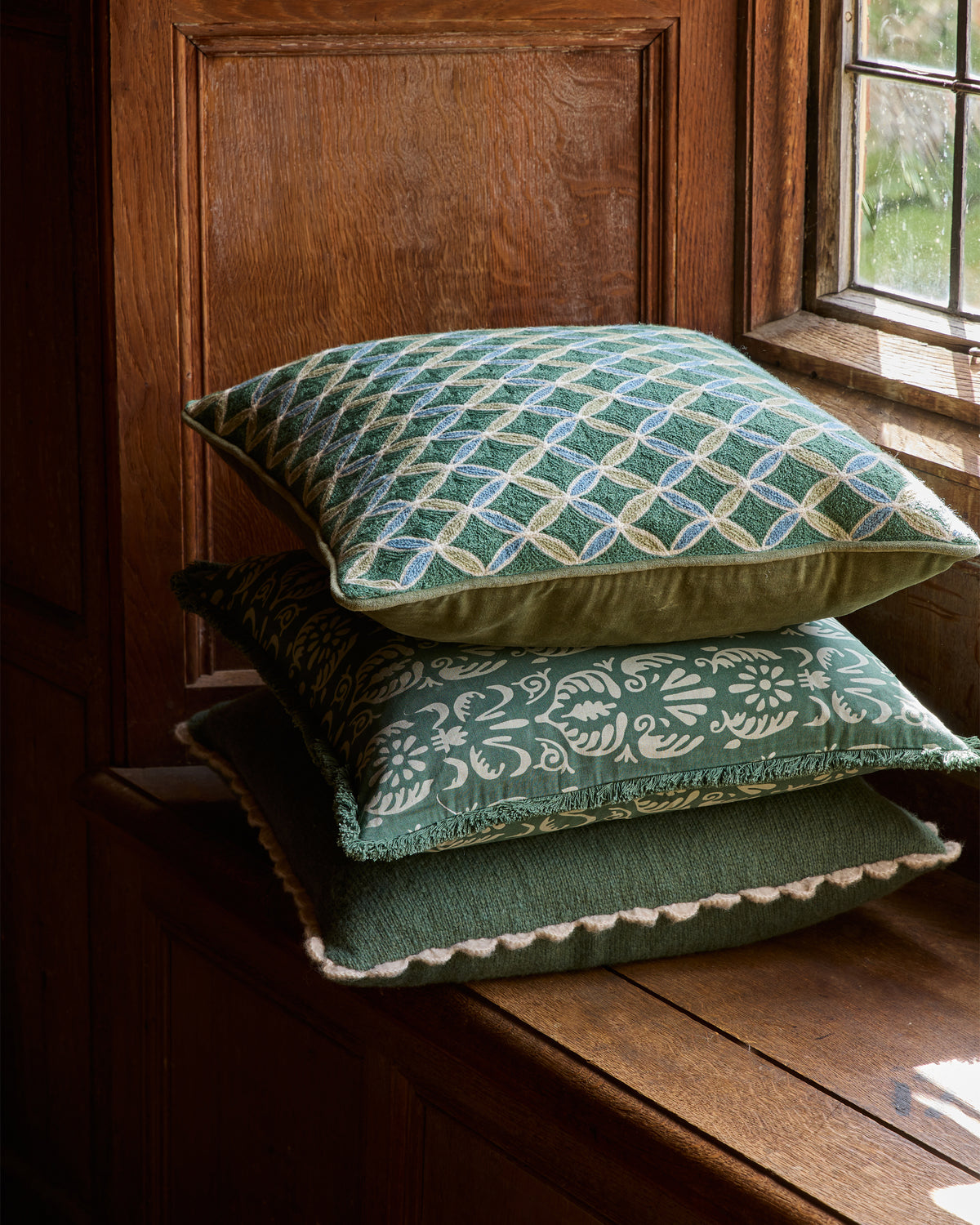 Three green patterned cushions on a wooden surface near a window.