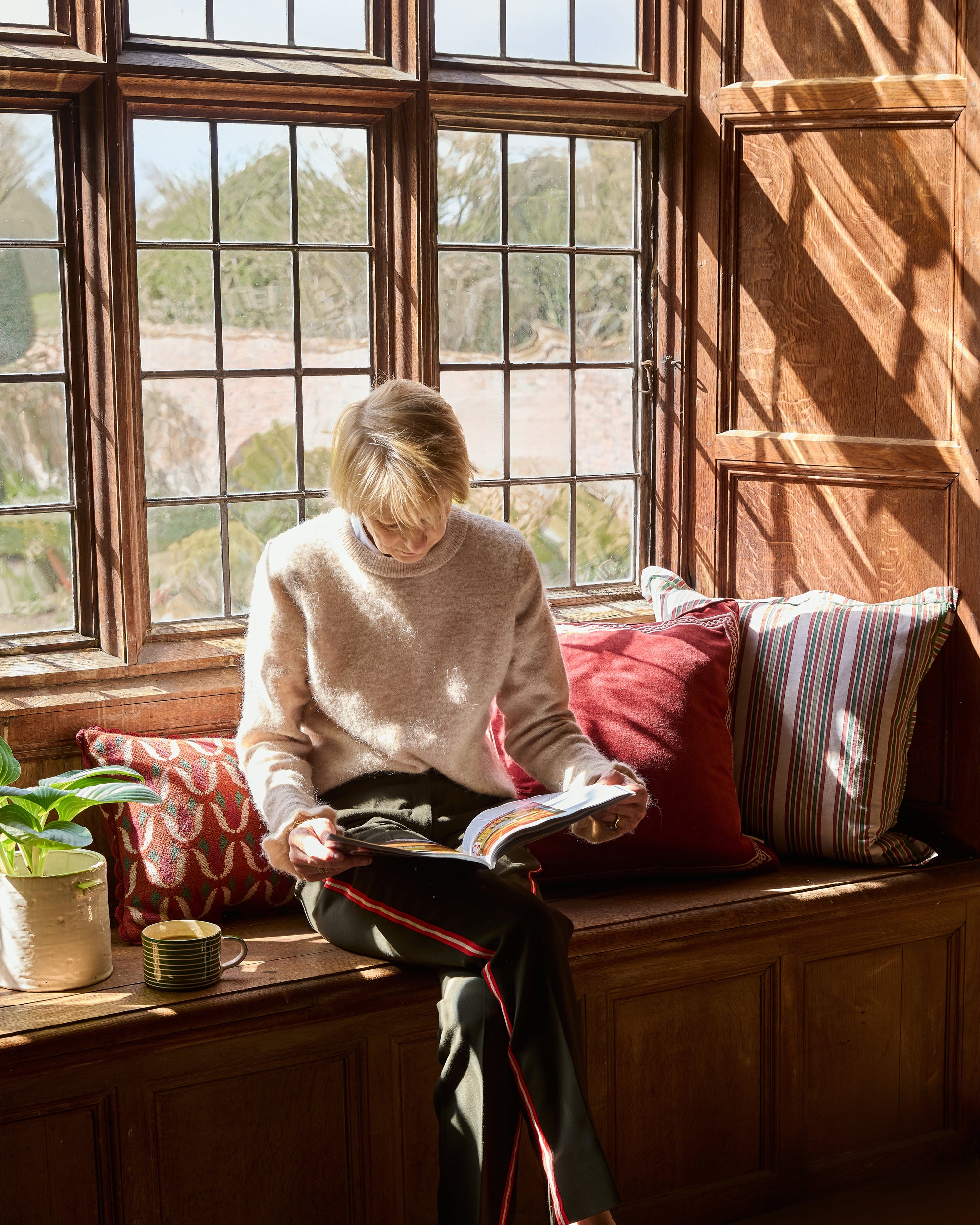 Person reading a book in a cozy room with large windows and wooden furniture.