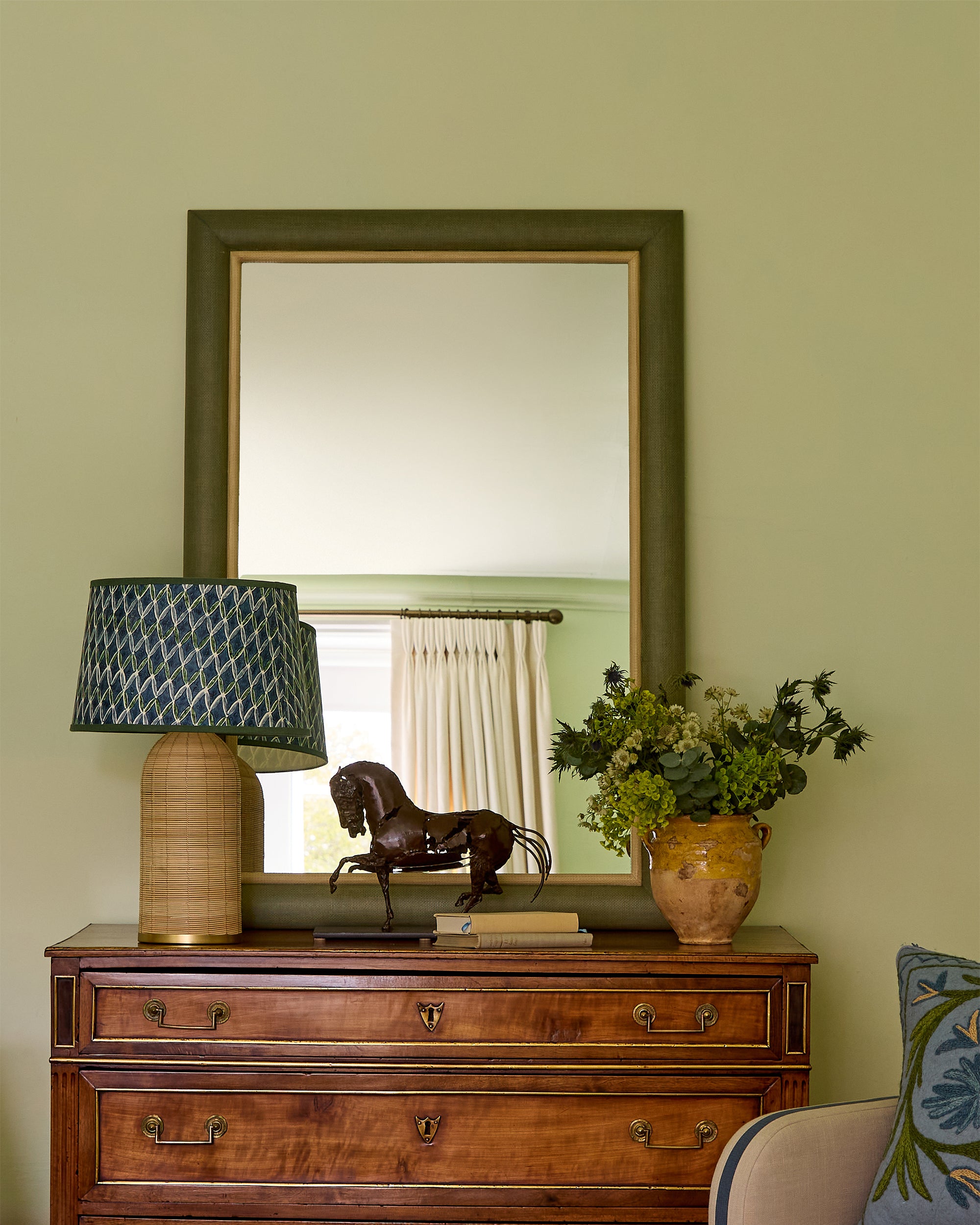 Wooden chest of drawers with mirror, lamp, and decorative items against a green wall.