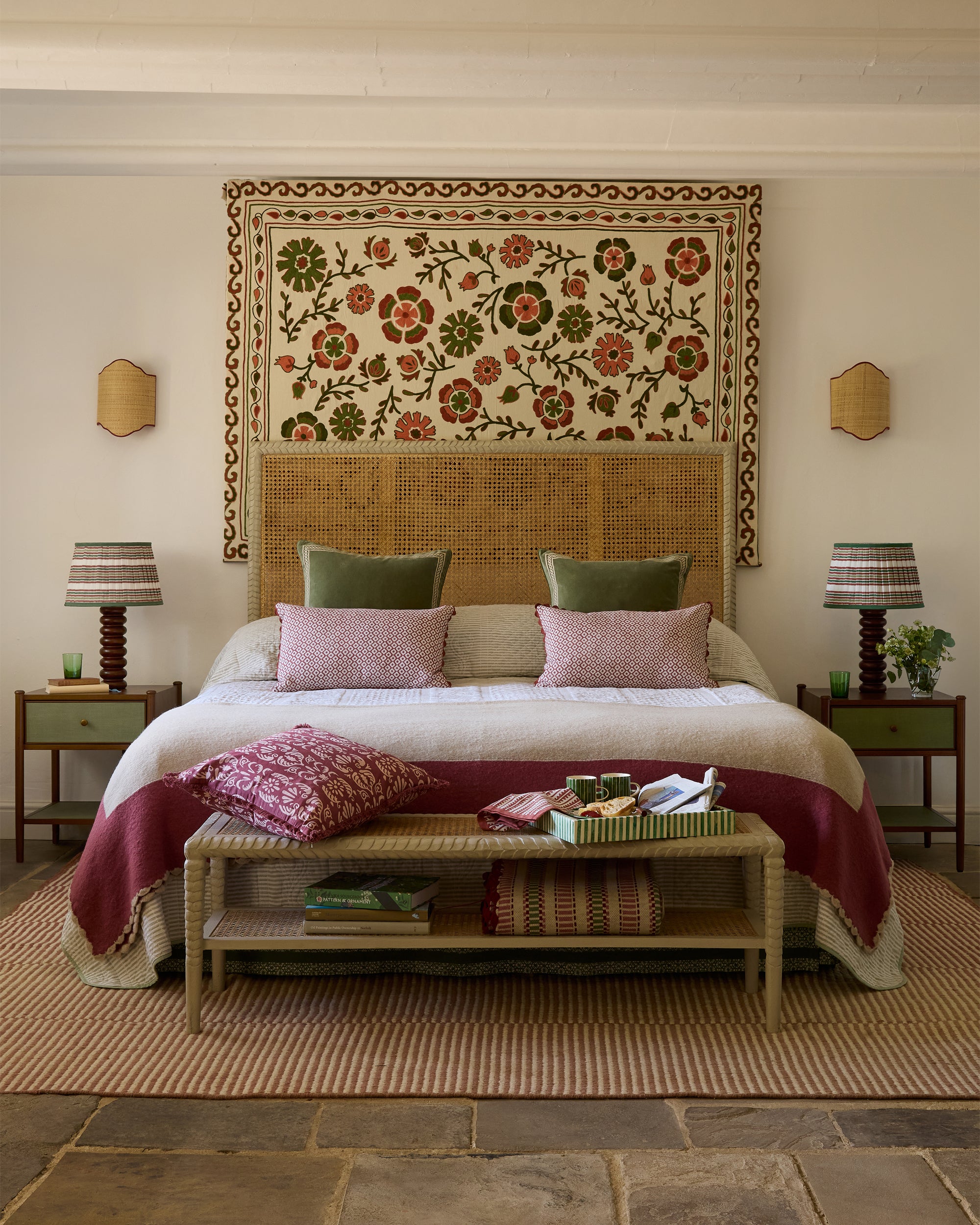 Decorative bedroom with a floral tapestry above a bed, two nightstands with lamps, and a woven bench.