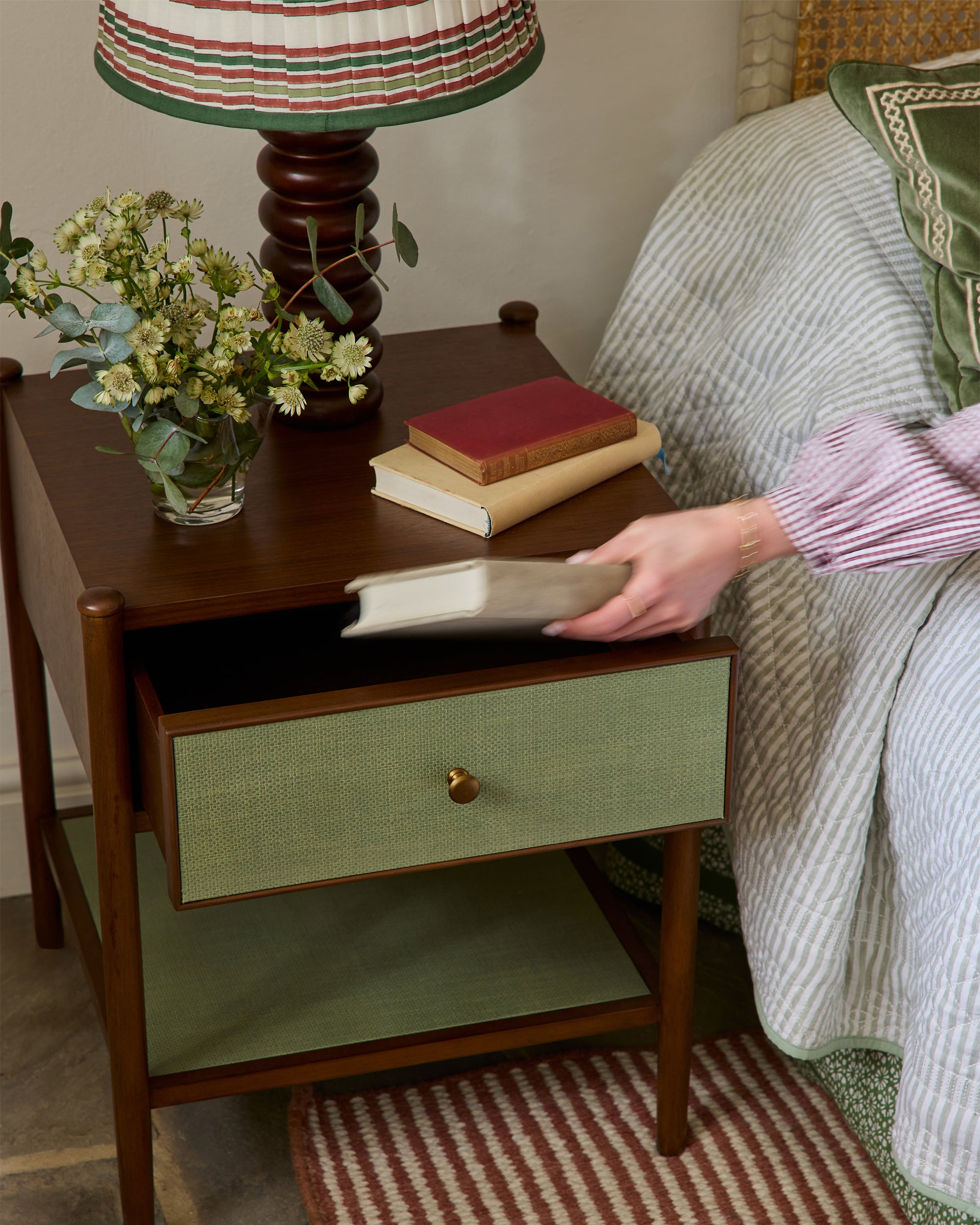 Person holding a book next to a wooden side table with a lamp and books on it.