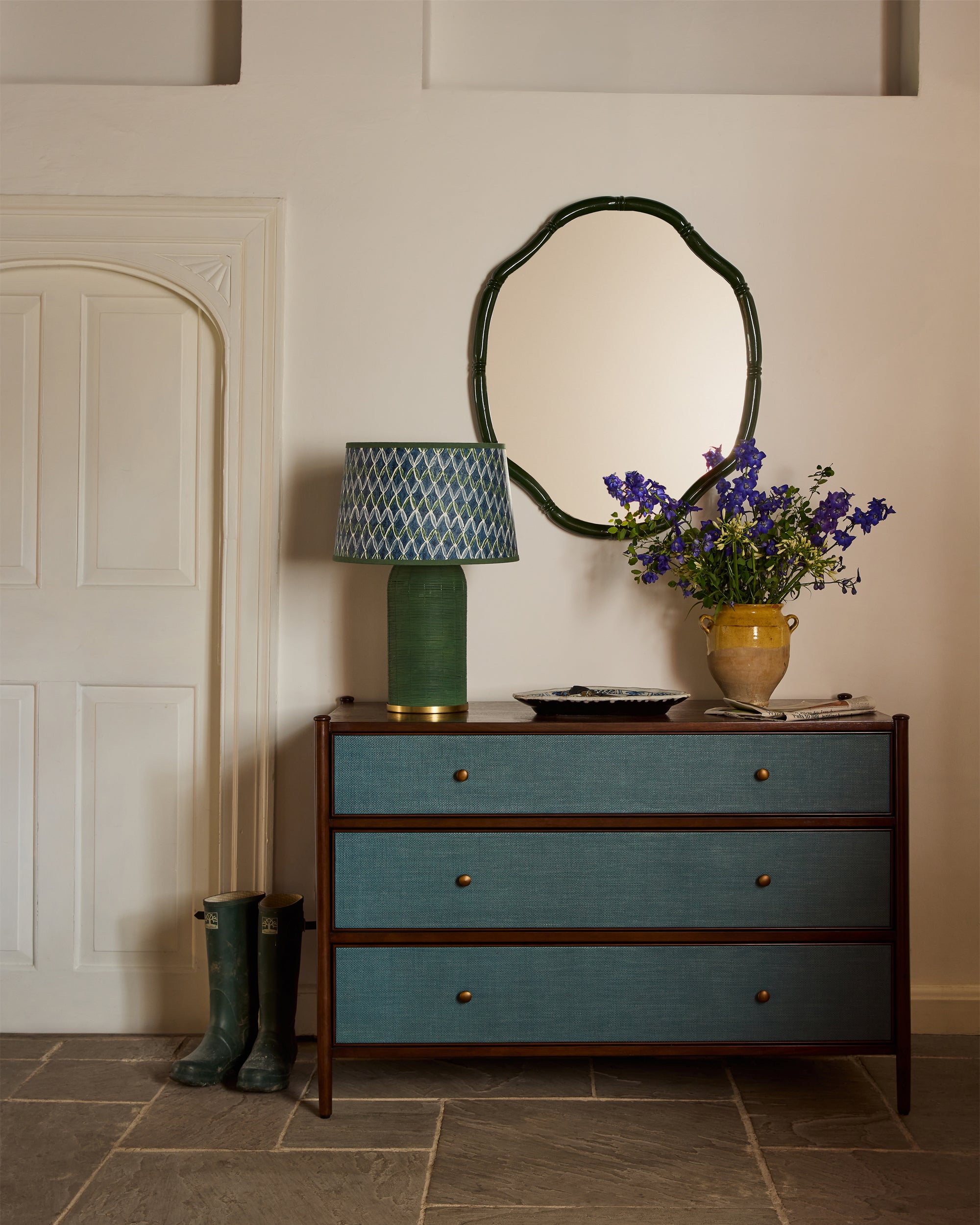 Blue chest of drawers with an oval mirror, green lamp, and vase of flowers against a white wall.