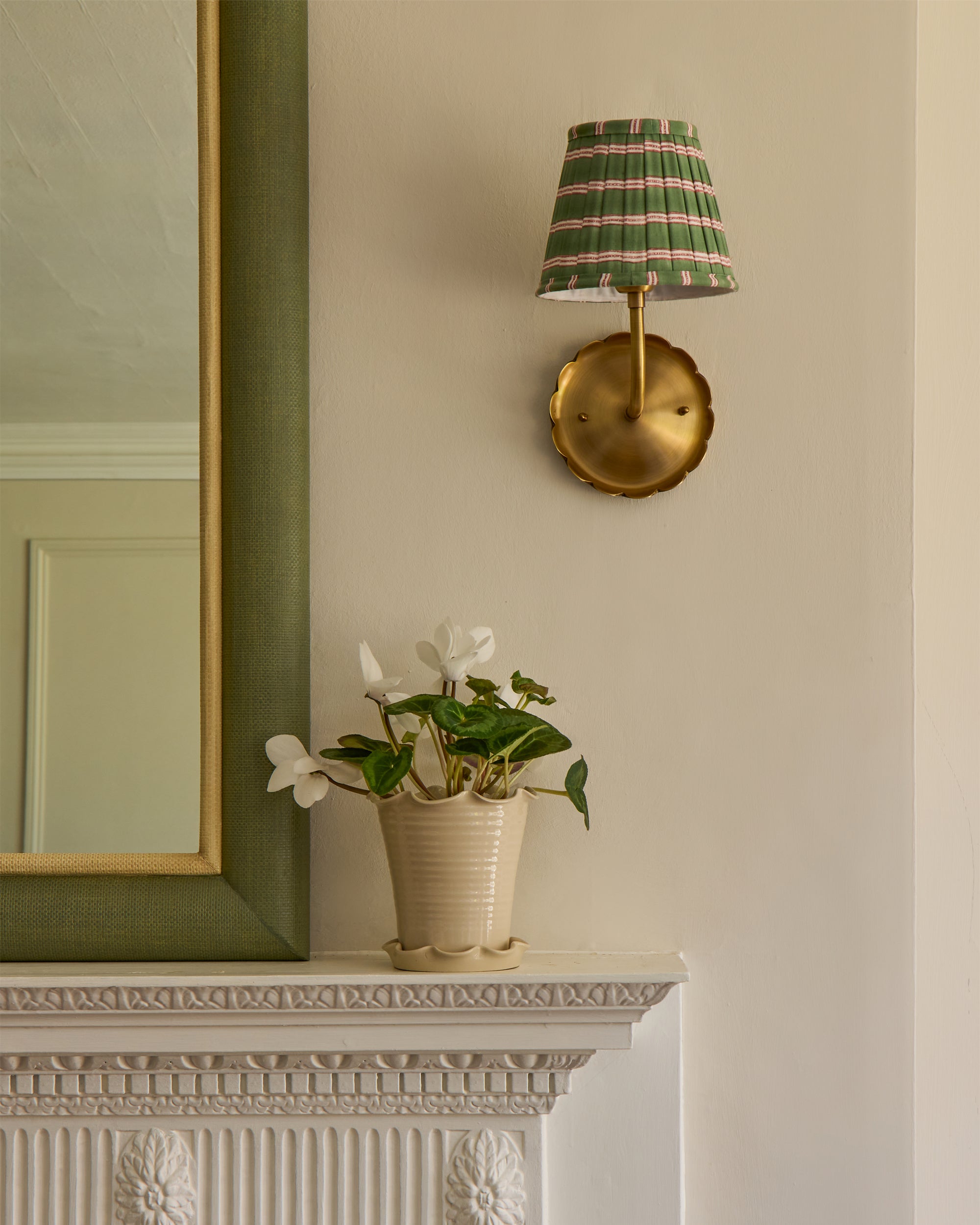 Brass wall light with striped edo shade above a potted plant on a fireplace mantle.