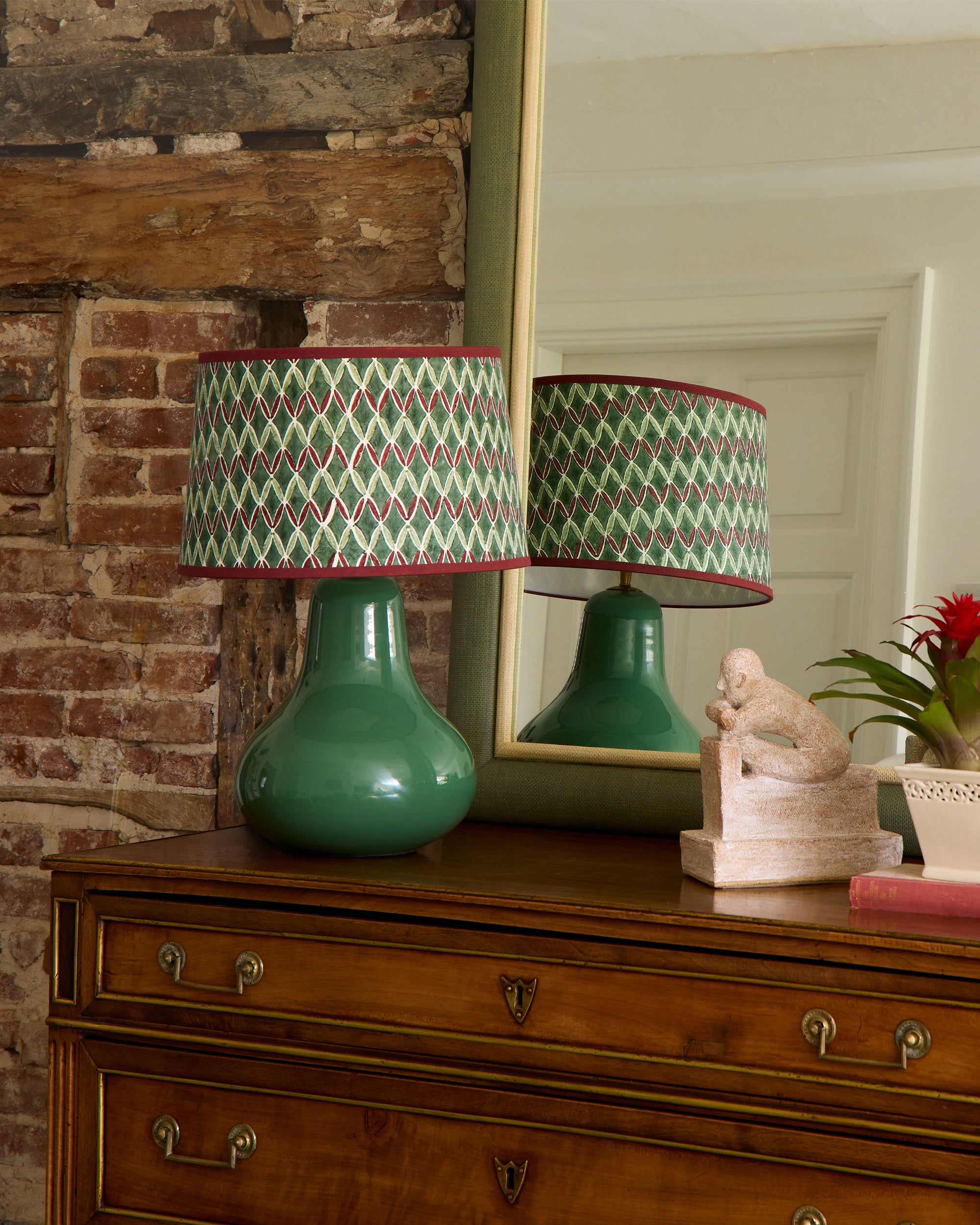 A green lamp with a patterned shade on a wooden dresser against a brick wall.