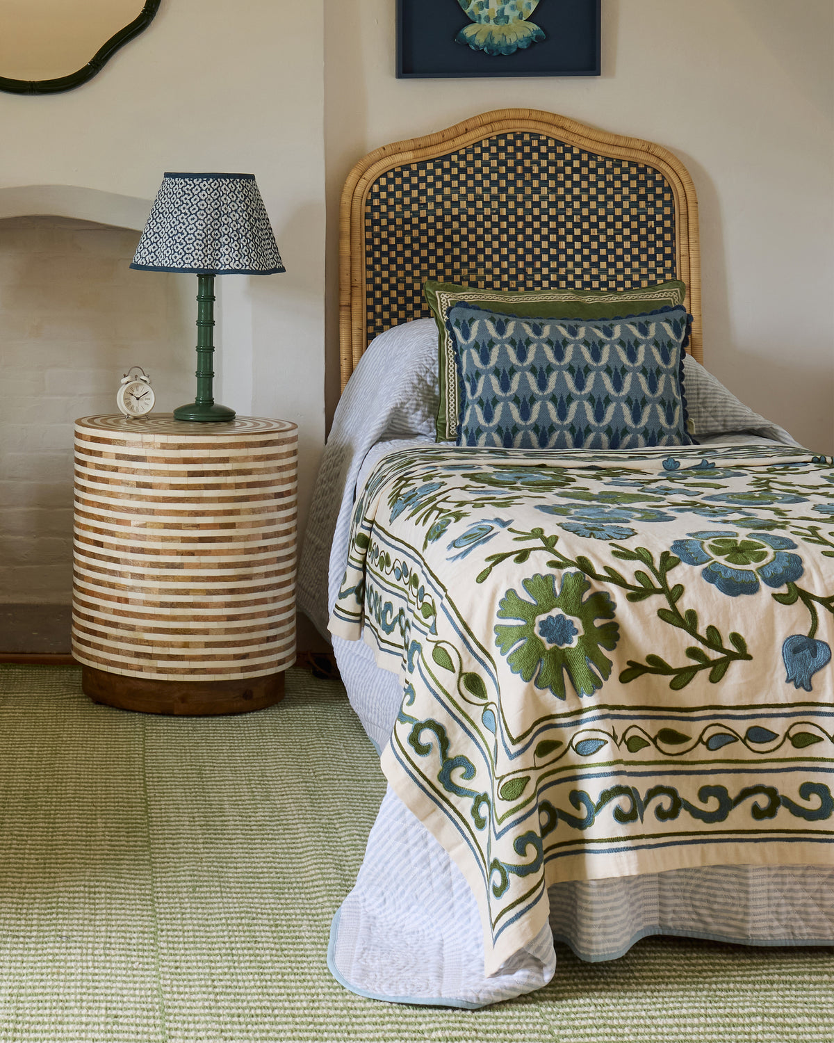A styled bedroom scene featuring a bed dressed in a cream quilt with green and blue floral embroidery, a patterned blue pillow, a woven rattan headboard, and a matching striped side table holding a blue patterned lamp.