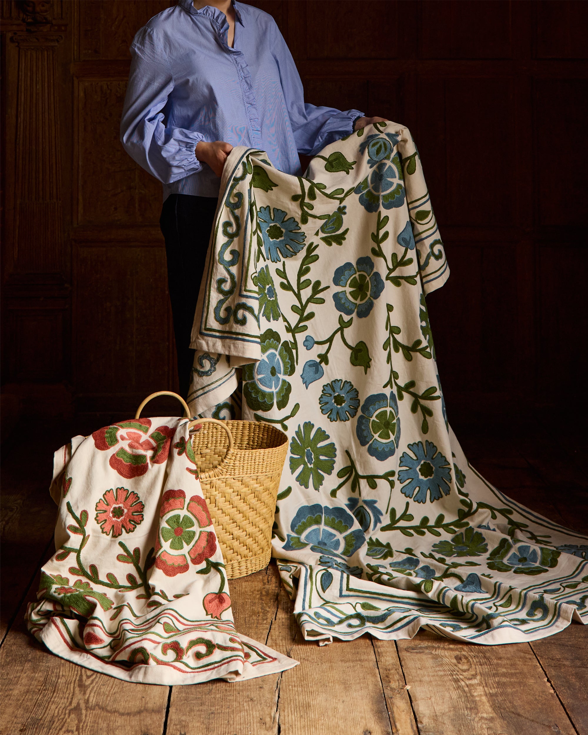 Person holding a floral-patterned suzani throw on a wooden floor with a dark background.