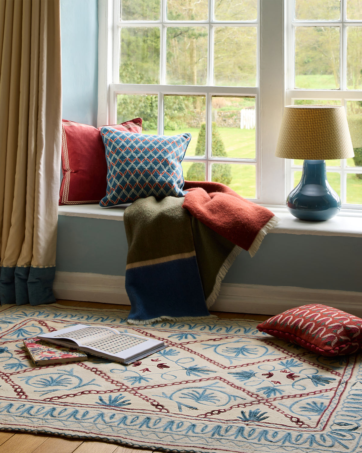 Cozy reading nook with cushions, a book, and a lamp in a room with large windows.