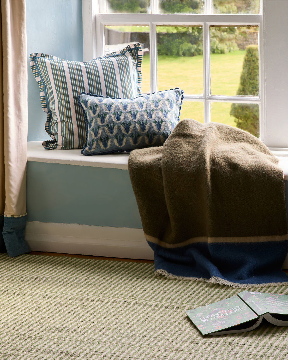 A cozy window seat setup with striped and patterned blue cushion, a brown and navy blanket draped over the sill, a light blue wall, and a textured green rug on the floor.