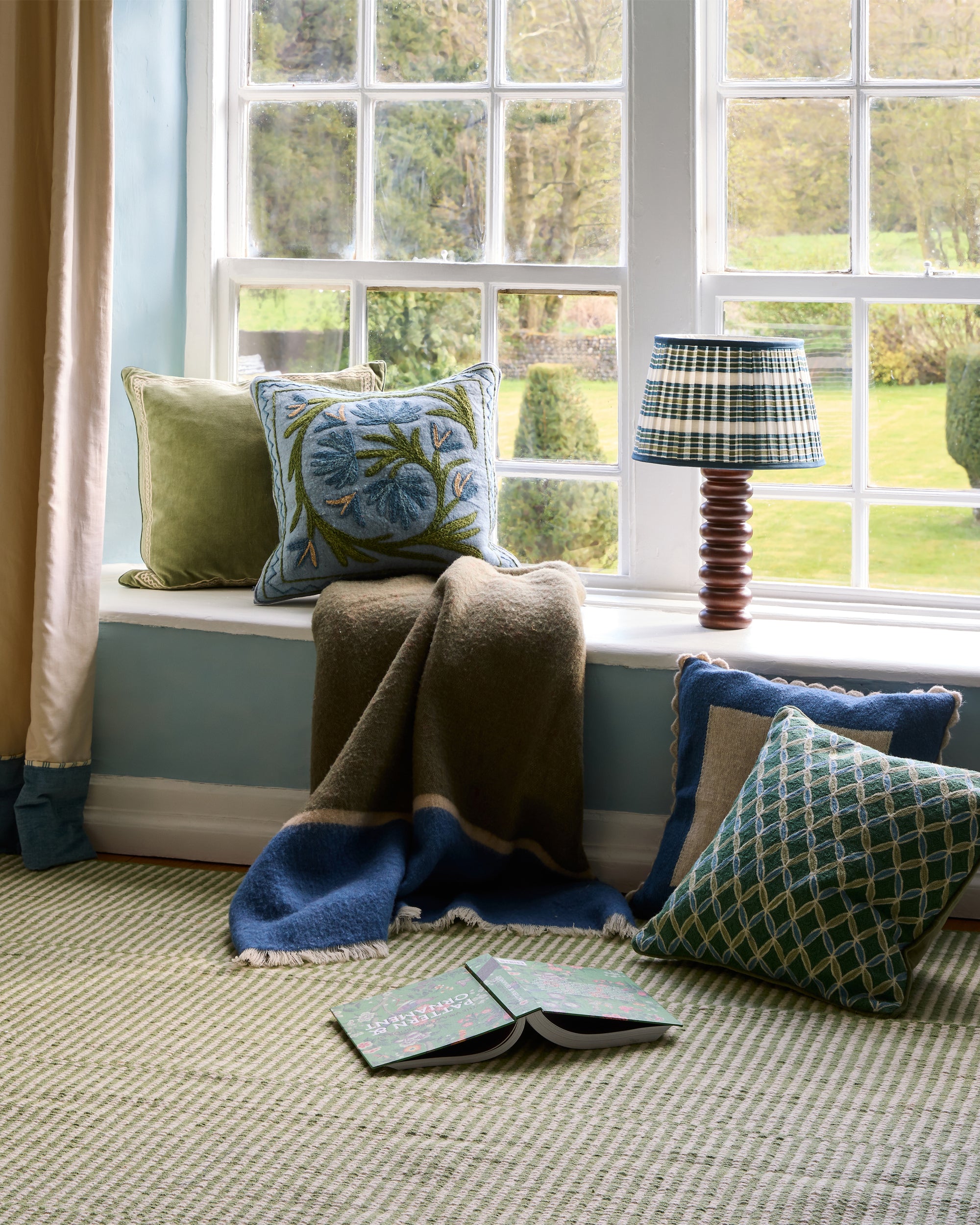 Cozy window seat with cushions, a blanket, and a lamp in a room with large windows.