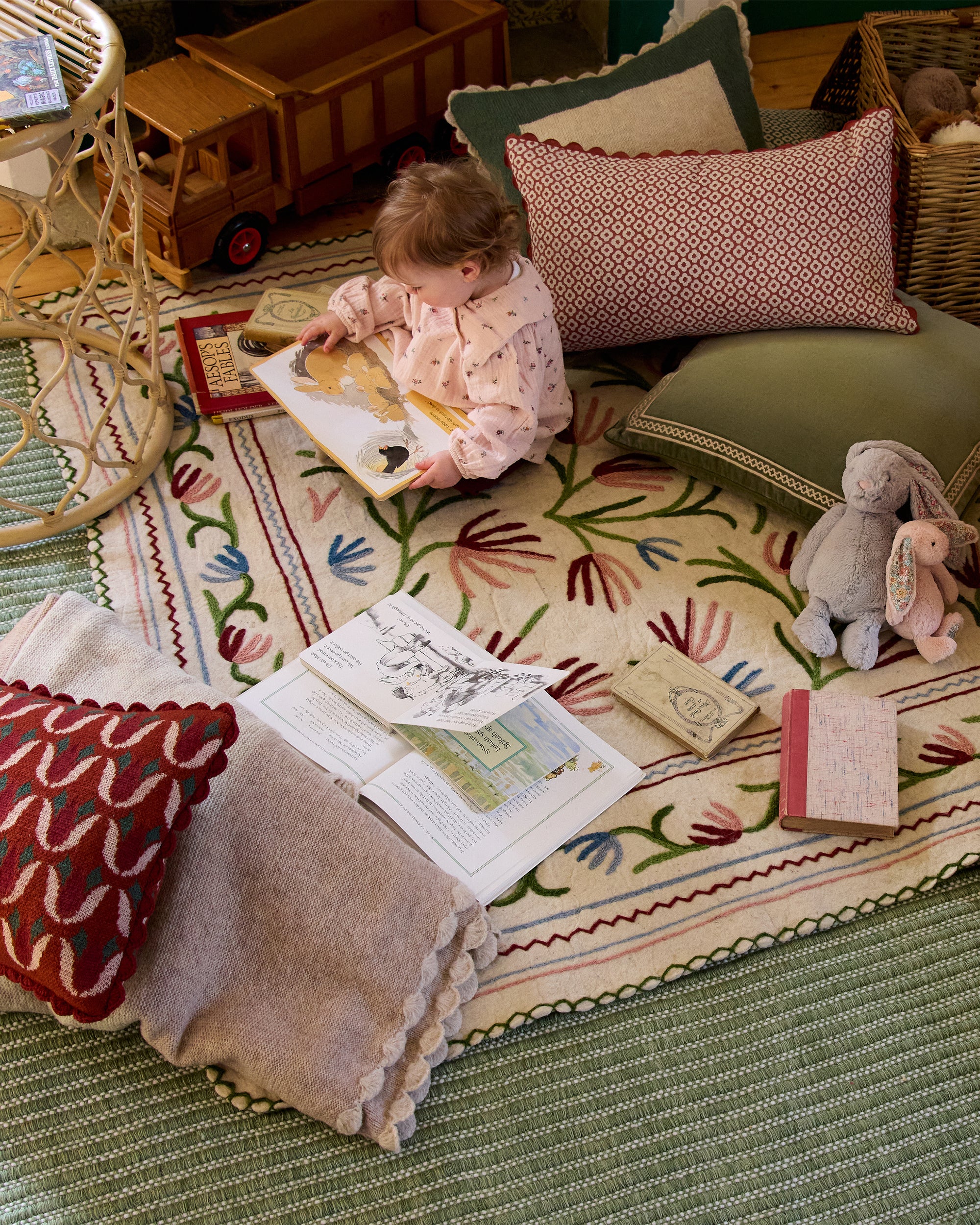 Child reading a book on a colourful rug with toys and cushions around.
