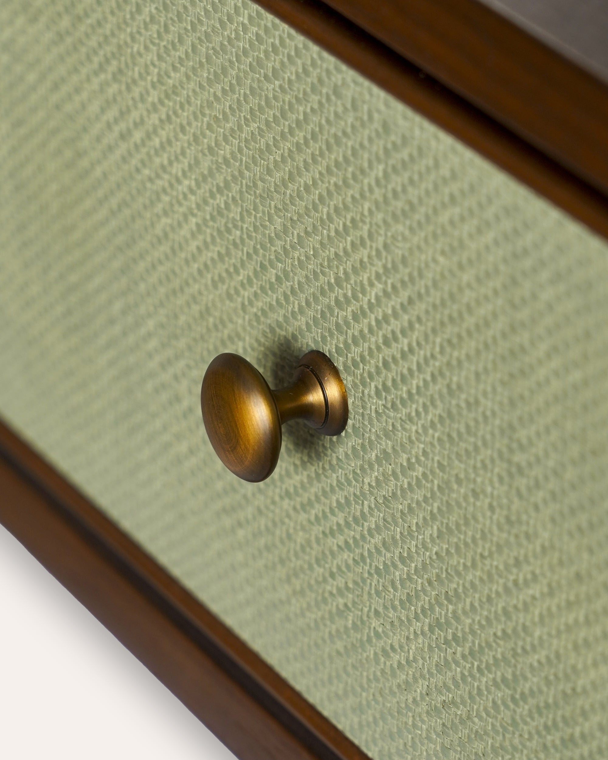 Close-up of a round brass drawer knob mounted on a light sage green textured drawer front with a dark wood frame.