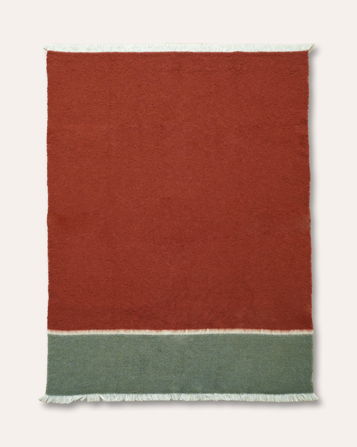A rectangular woven throw blanket with a solid rust-red upper section, a muted sage green lower band, and white fringe on all sides, shown flat against a light cream background.