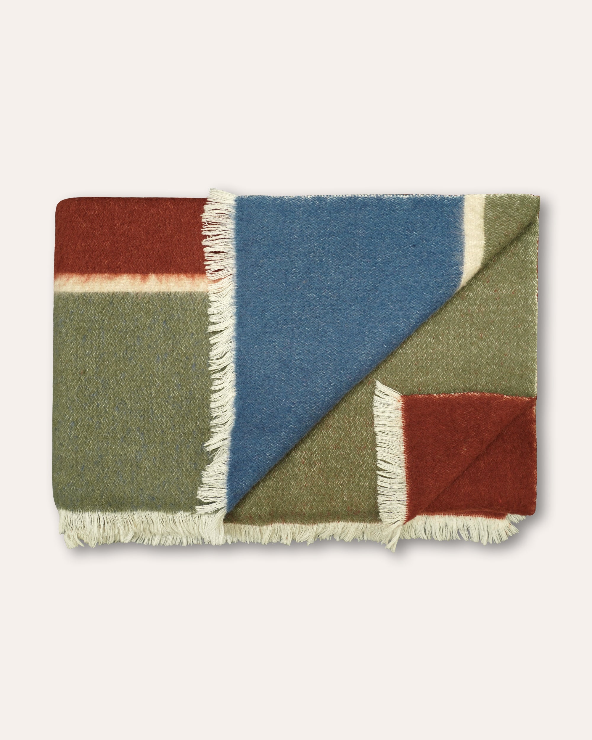 A folded woven throw blanket with a geometric patchwork design in muted blue, olive green, rust red, and cream, finished with white fringe on all sides, shown against a light beige background.
