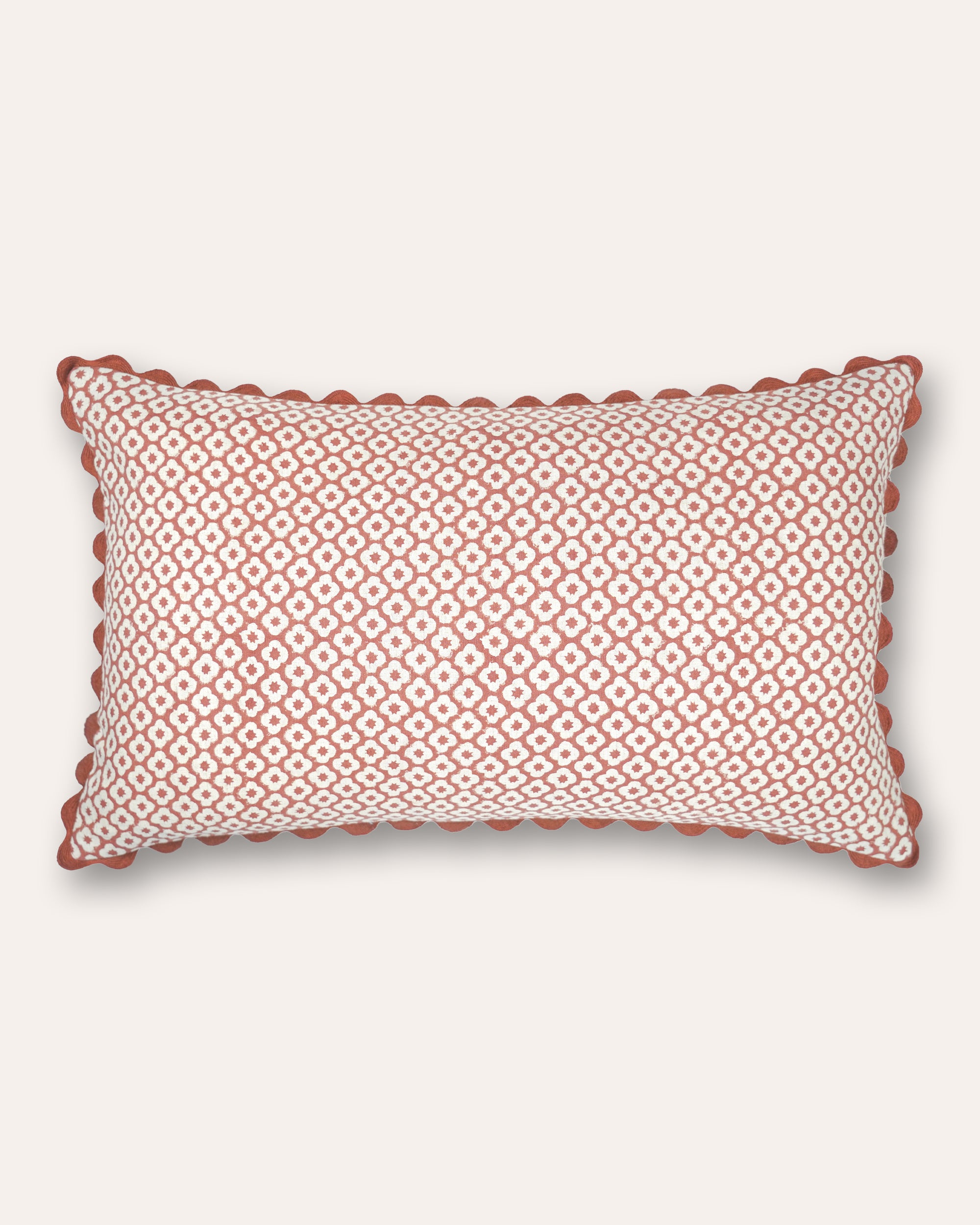 A rectangular cushion featuring a repeating geometric diamond pattern in terracotta and off-white, finished with a scalloped terracotta border, shown against a light cream background.