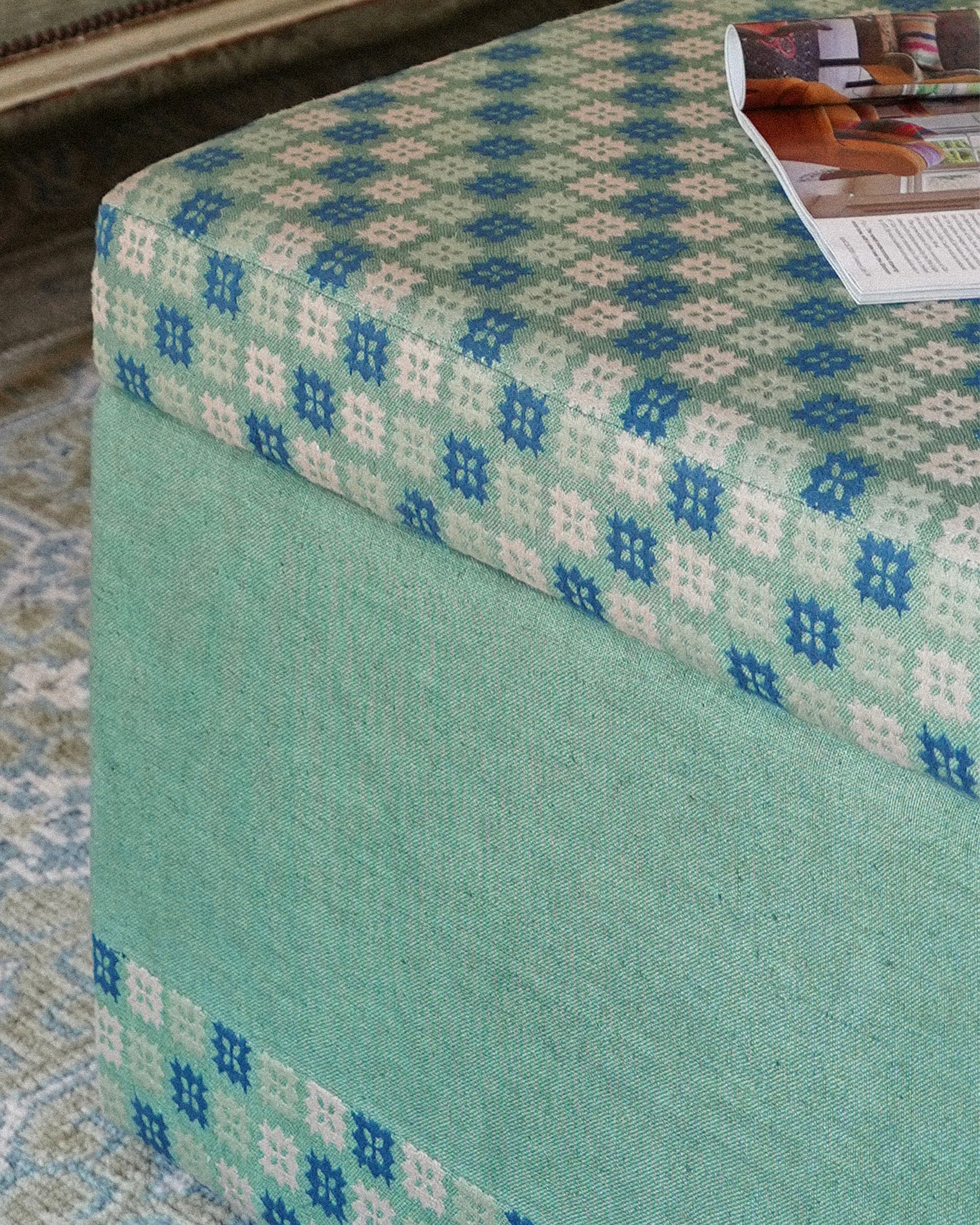 A close-up of a upholstered ottoman or bench corner, showing a mint green fabric base and a patterned top cushion in blue, sage green, and cream. A magazine rests on the cushion, and part of a patterned rug is visible underneath.