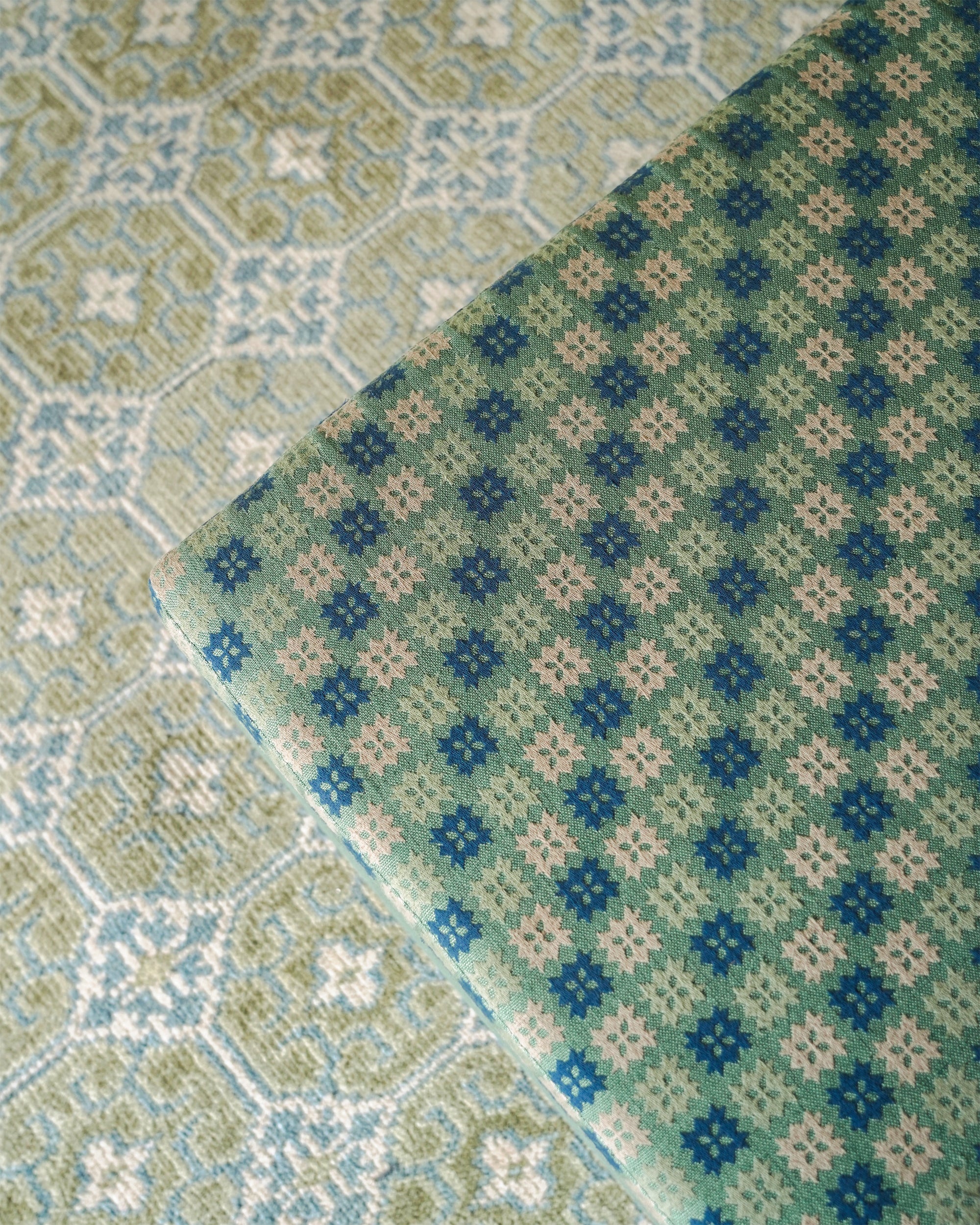 A close-up of a folded woven textile with a repeating geometric diamond pattern in blue, sage green, and cream, layered over a similar patterned fabric background.