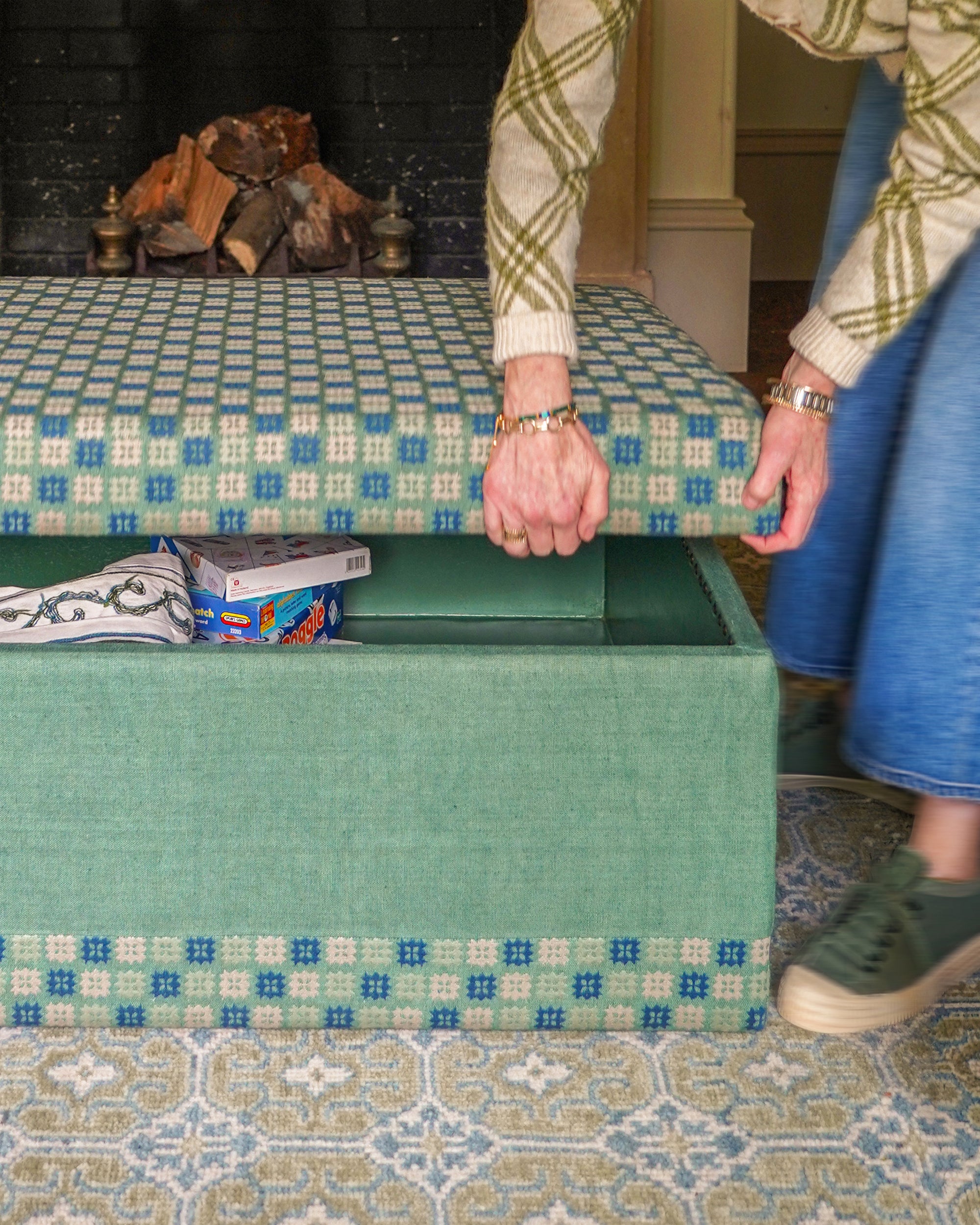 A person lifts the lid of a green storage ottoman in a cozy living room setting, revealing a shelf with books and a shoe box underneath. The ottoman features a sage green fabric body with a blue and cream checkered pattern along the top and bottom edge.