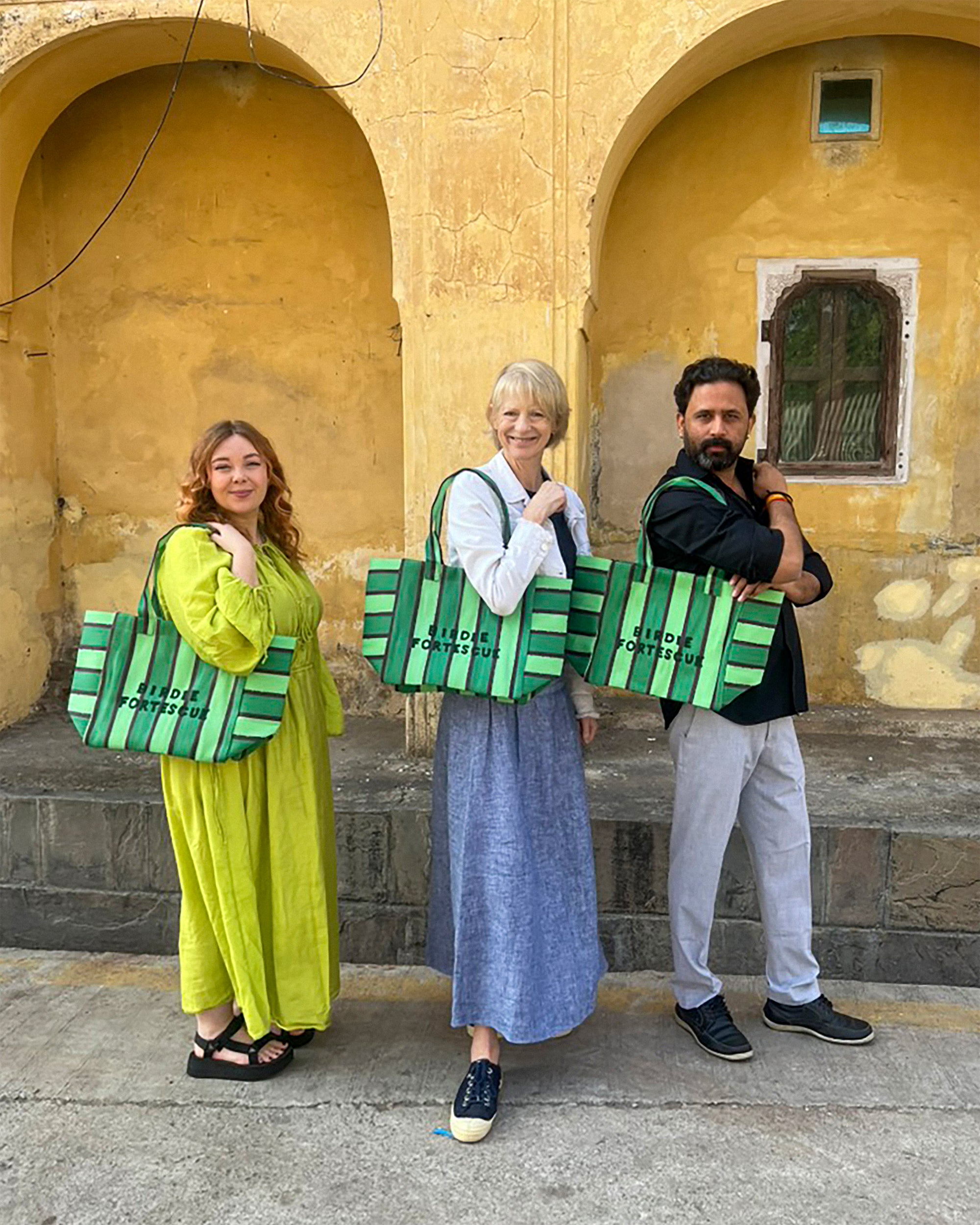 Three people holding green and black patterned bags in front of a yellow wall with arches.