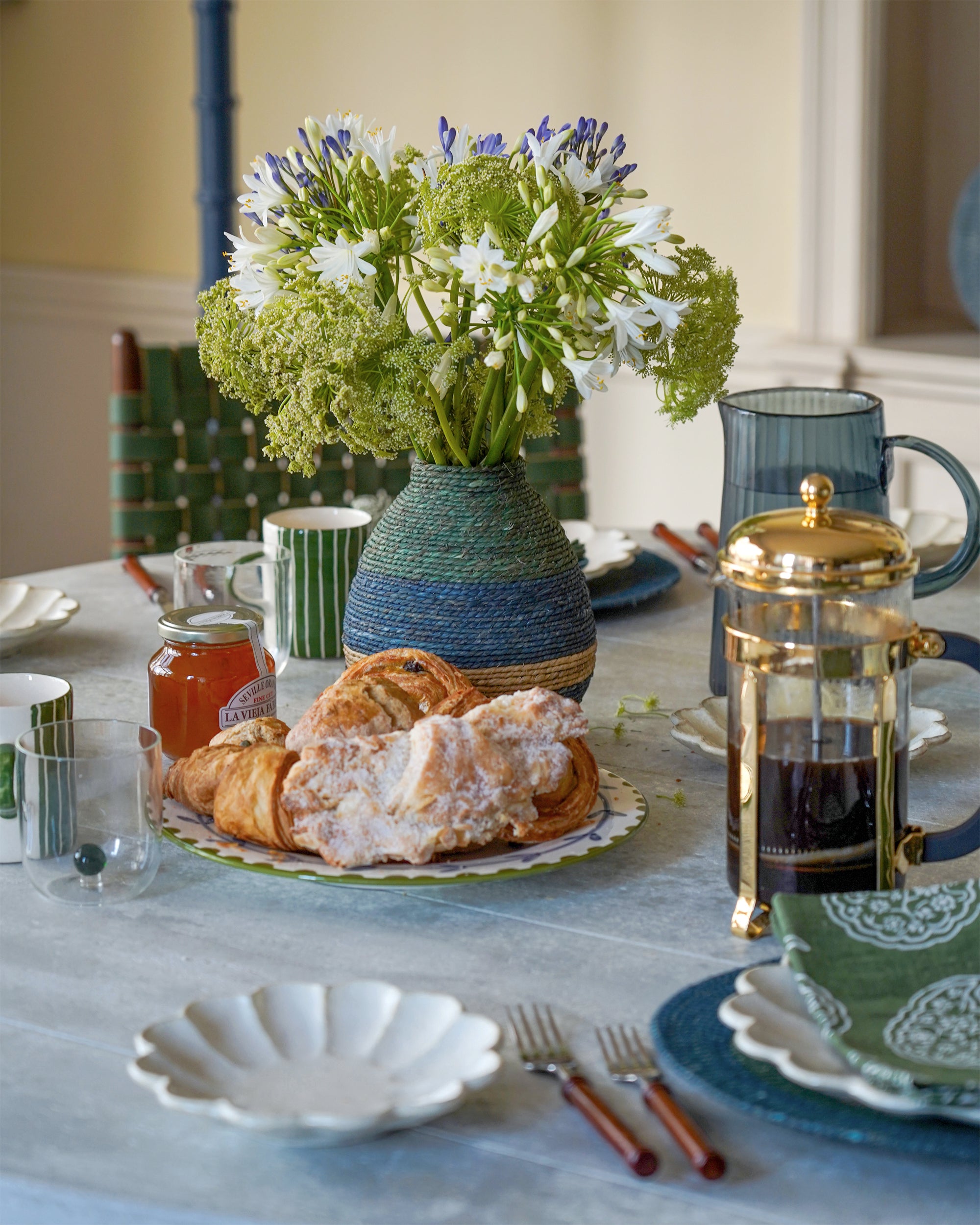 Table setting with pastries, flowers, and a French press on a light blue tablecloth.