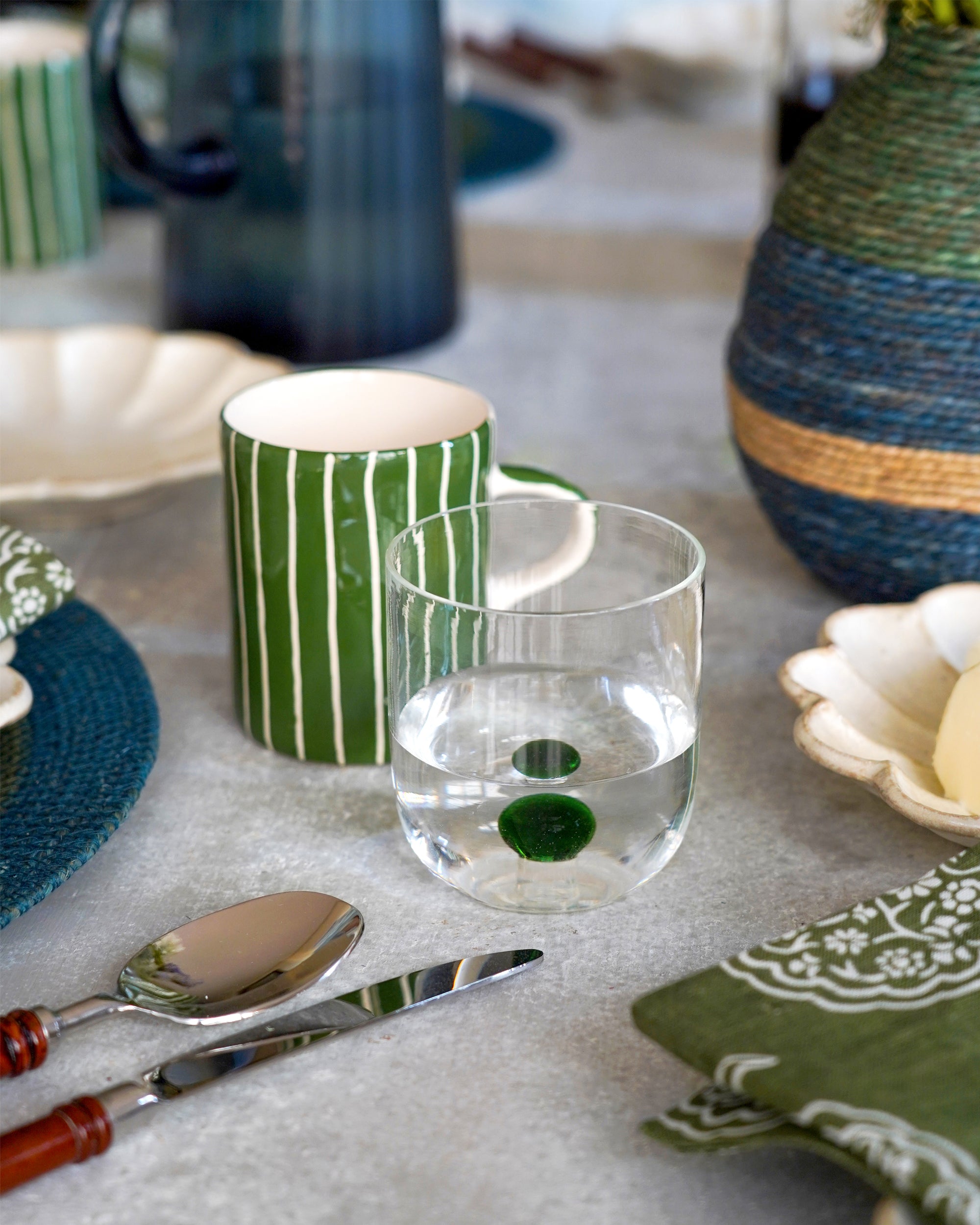 Table setting with a glass of water, green striped mug, and decorative items on a light surface.