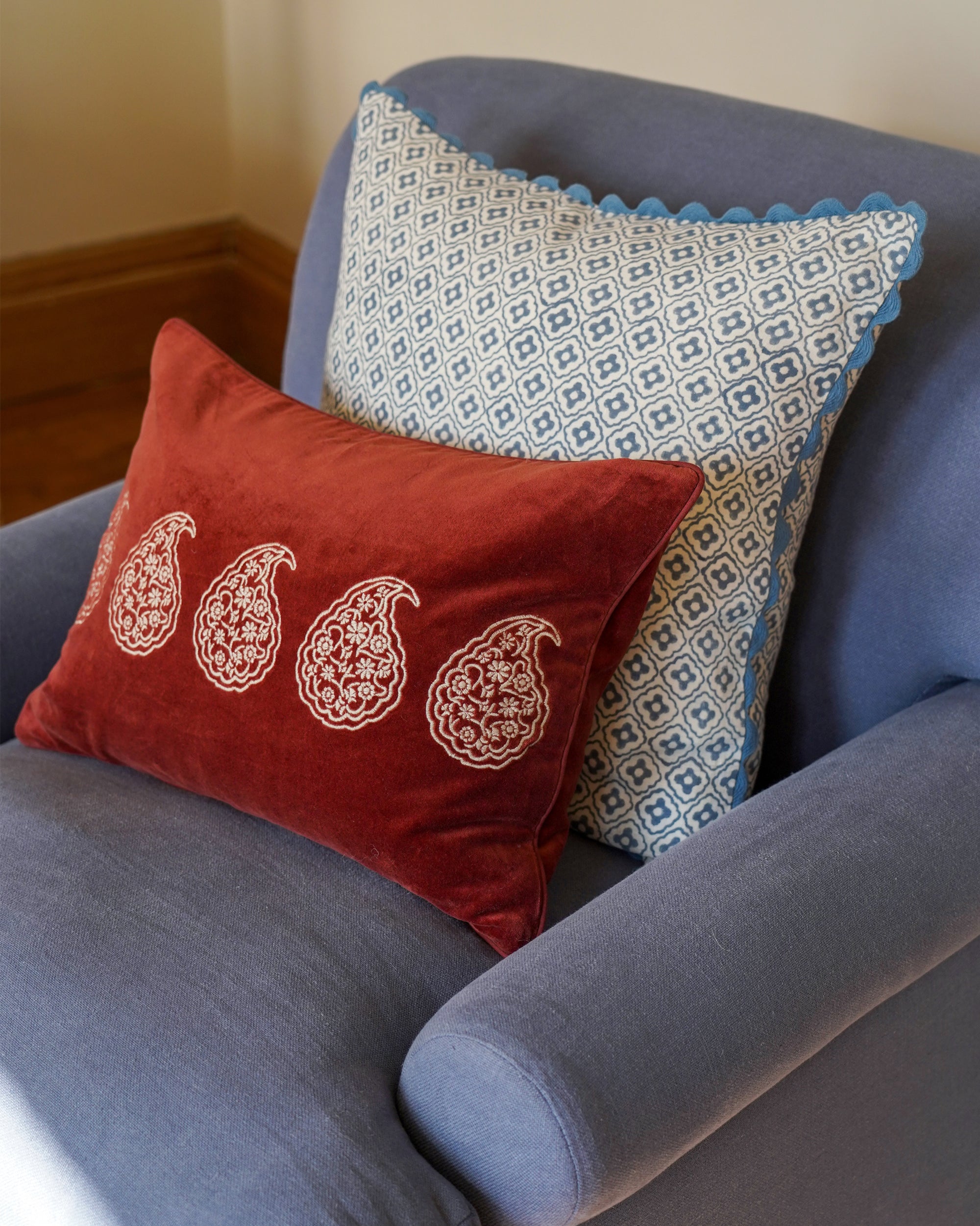 Two decorative pillows on a blue armchair with a red cushion featuring paisley patterns and a white pillow with blue trim.