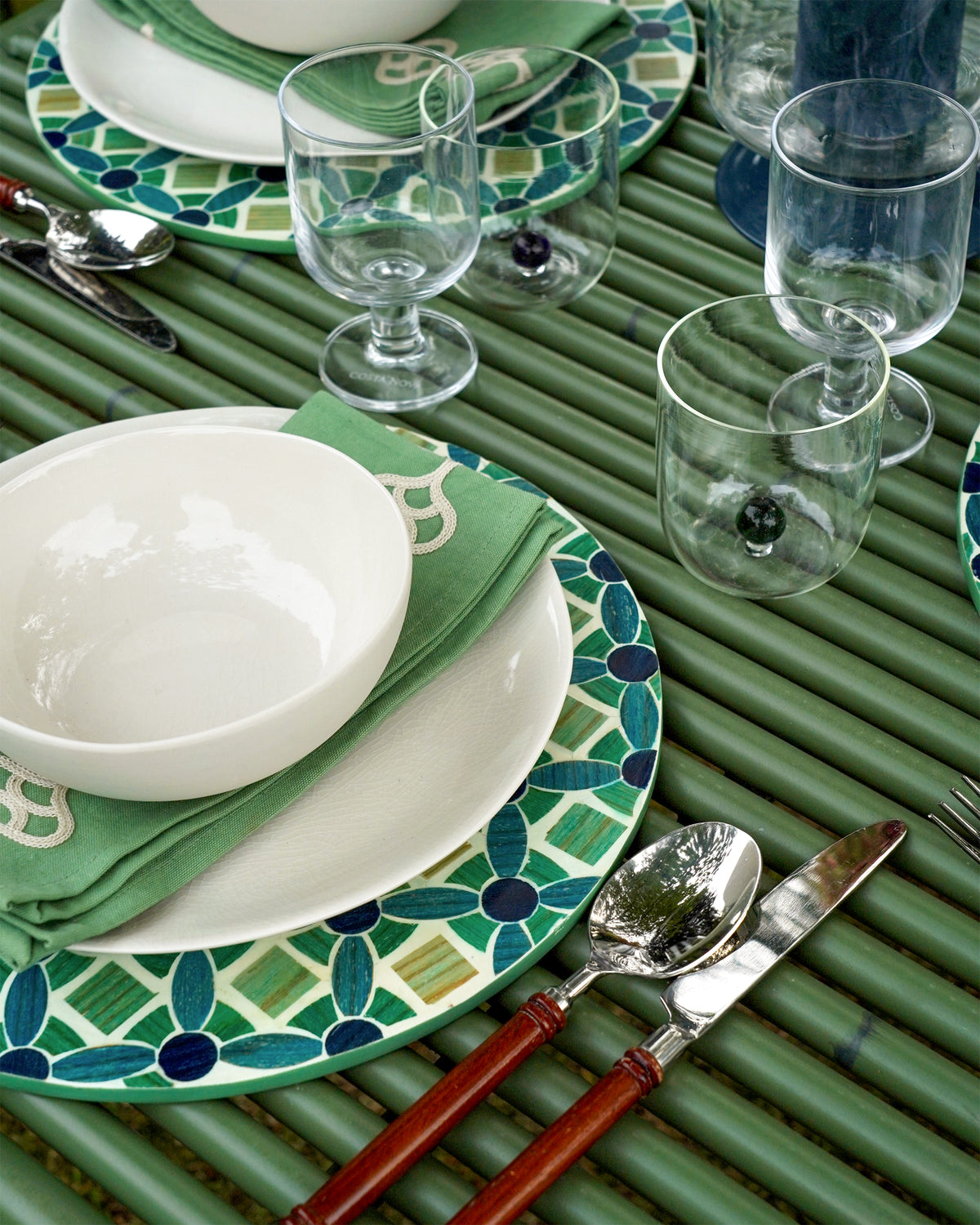 Dining table setting with green and white plates, glasses, and cutlery on a green placemat.