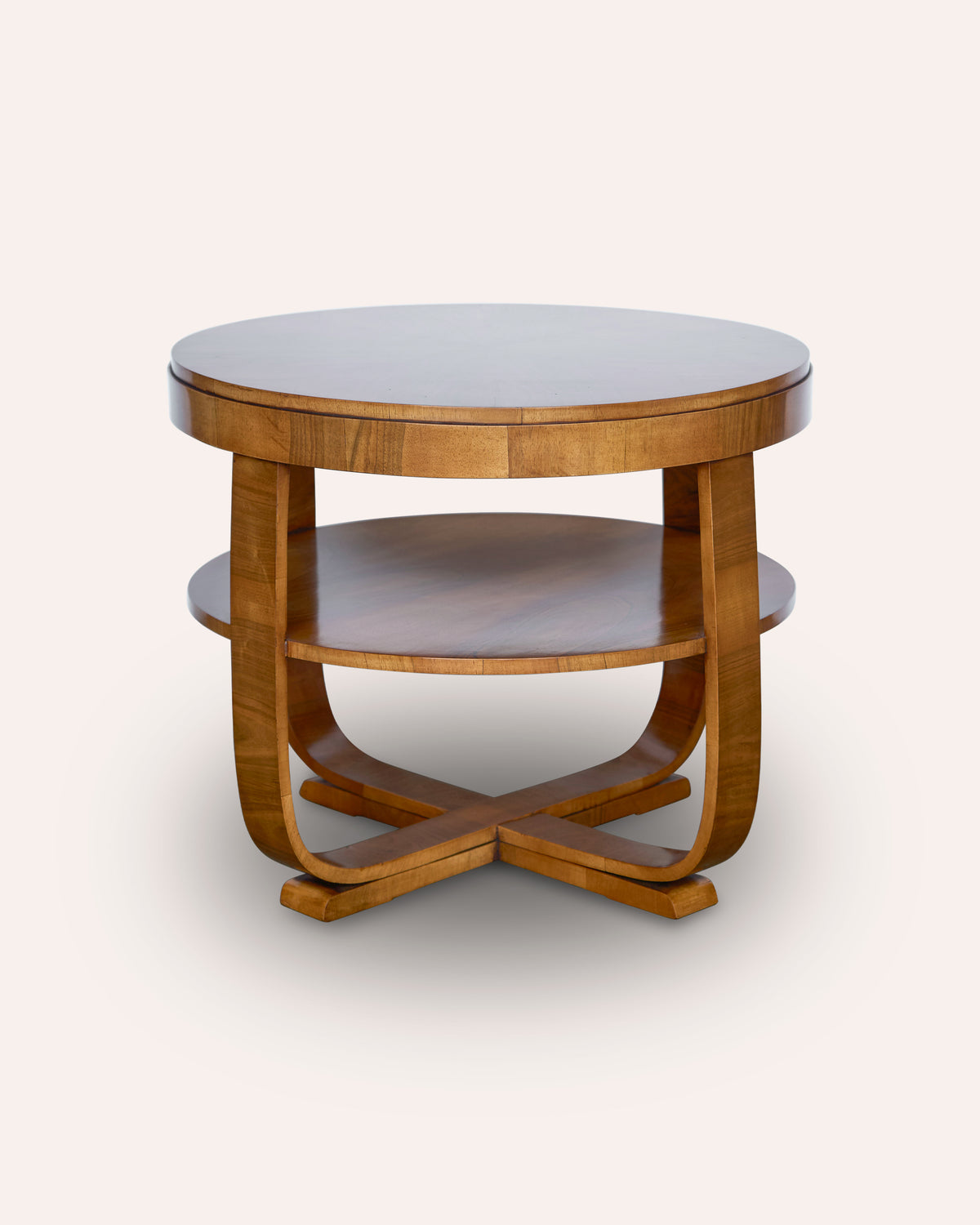 A mid-century modern round wooden end table with a warm medium-brown finish, featuring a top surface, a lower shelf, and curved legs that form an X base, set against a plain off-white background.