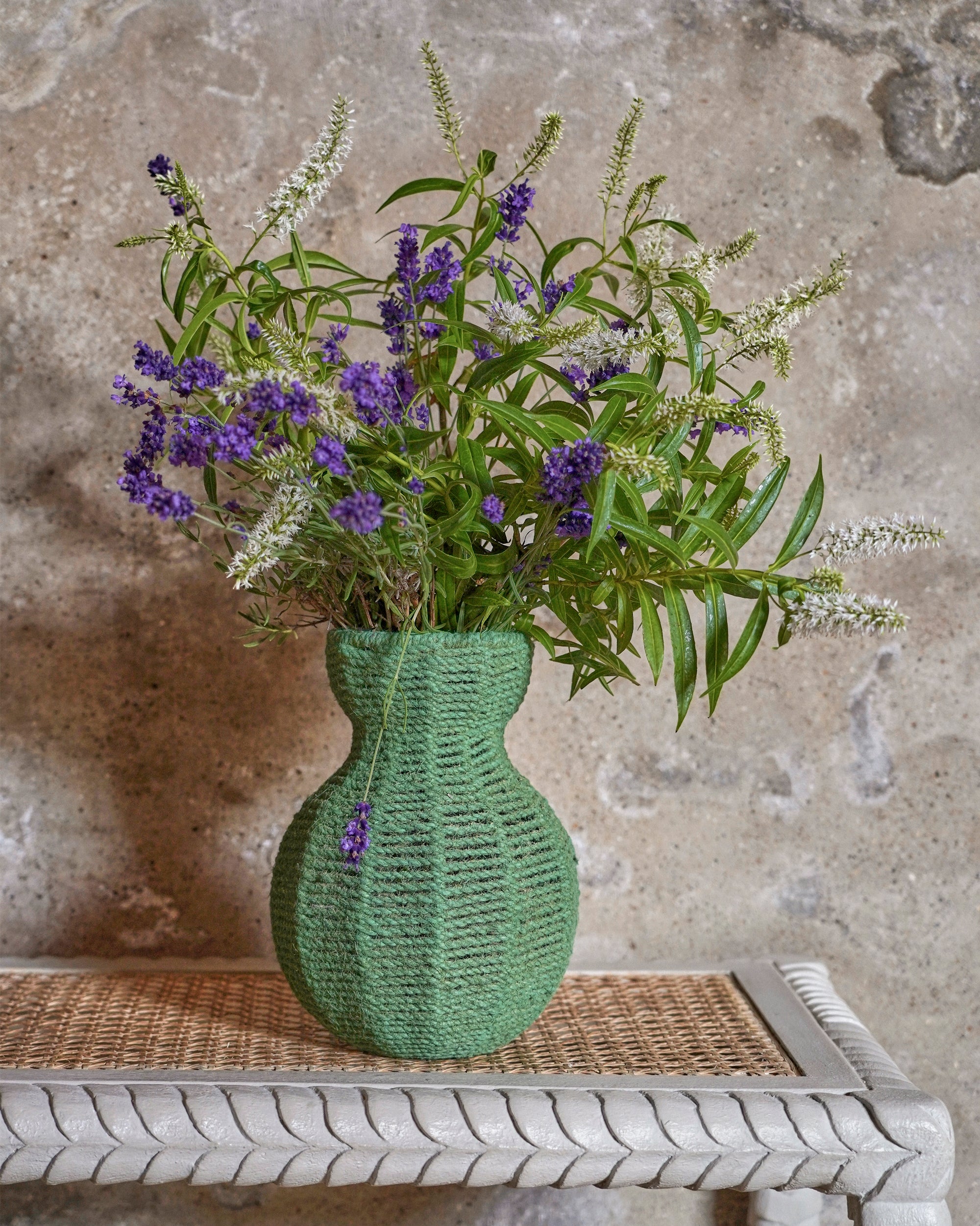 Green woven vase with purple and white flowers on a textured stone surface