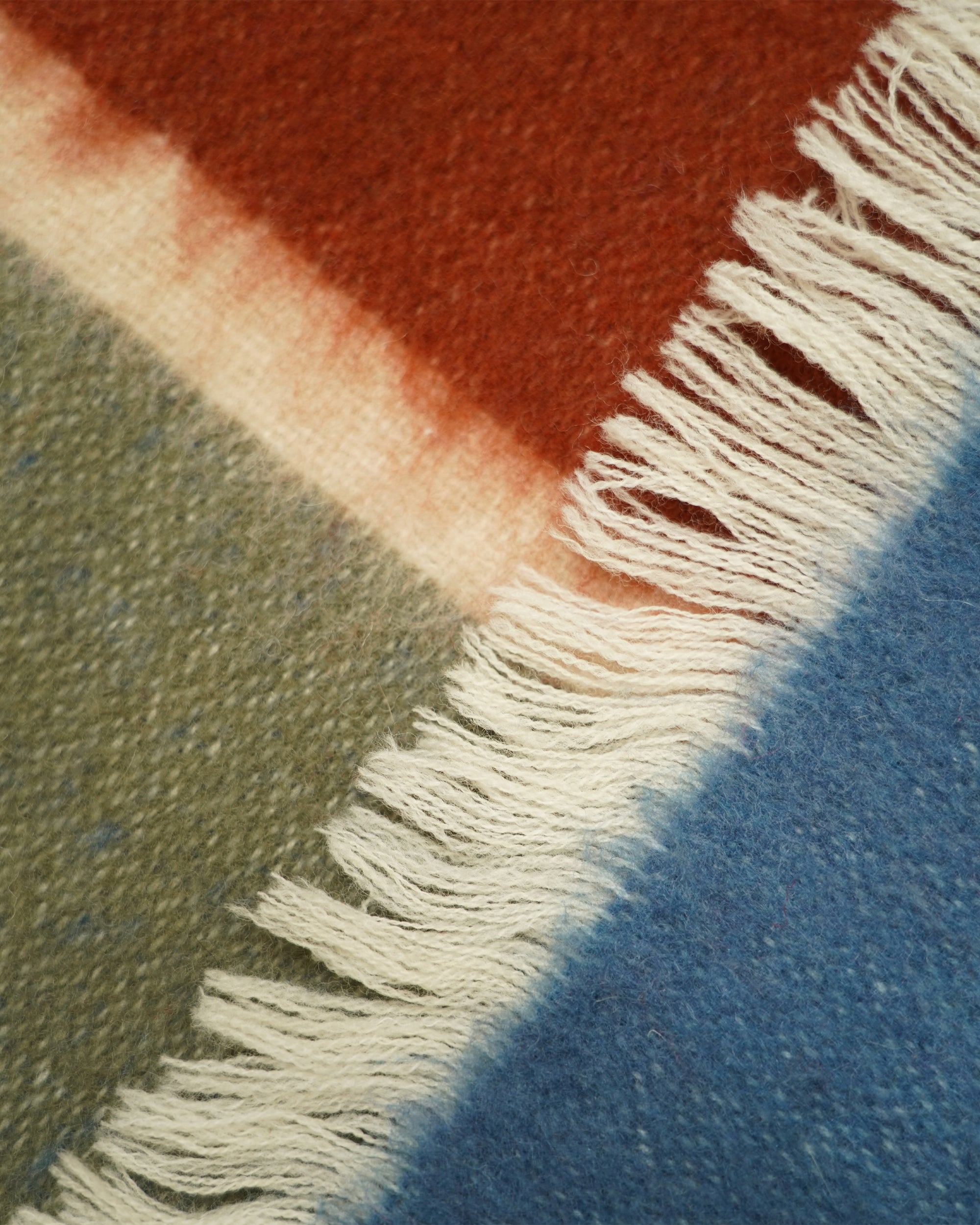 Close-up of a woven textile with fringe, showing broad color blocks in rust red, cream, olive green, and blue.