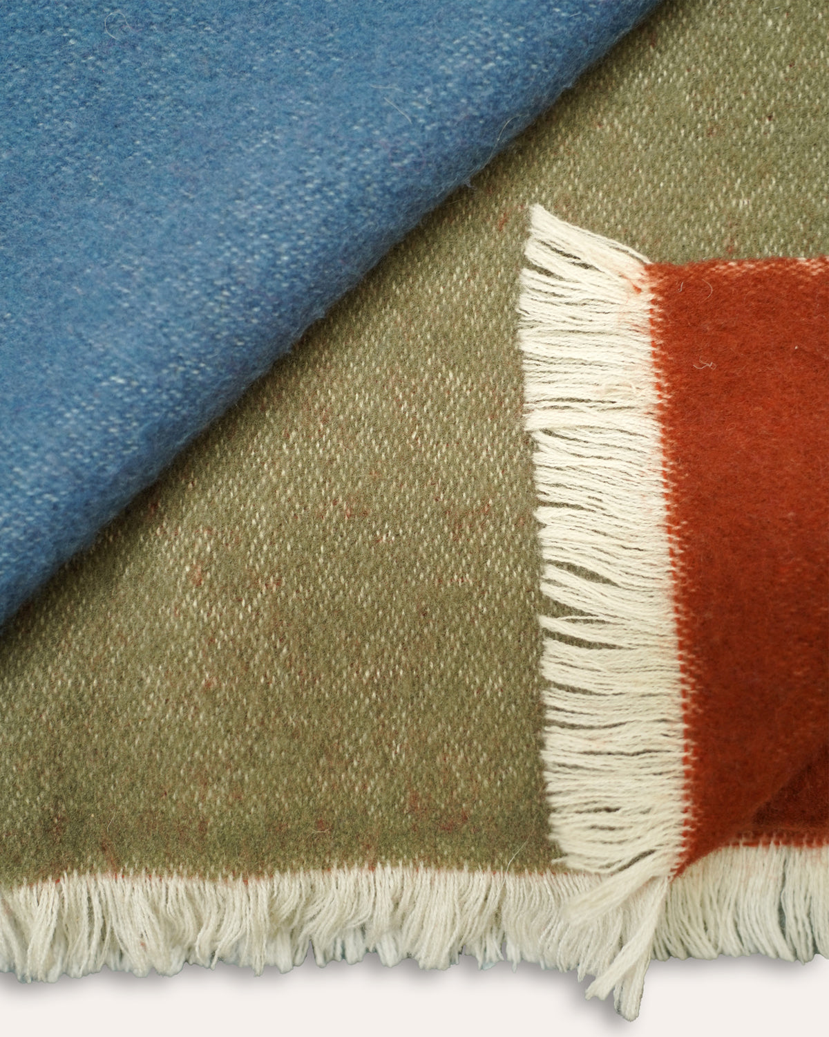 A close-up of a woven textile with fringe, showing a muted olive green main field, a rust orange block, and a blue section folded over at the top left.