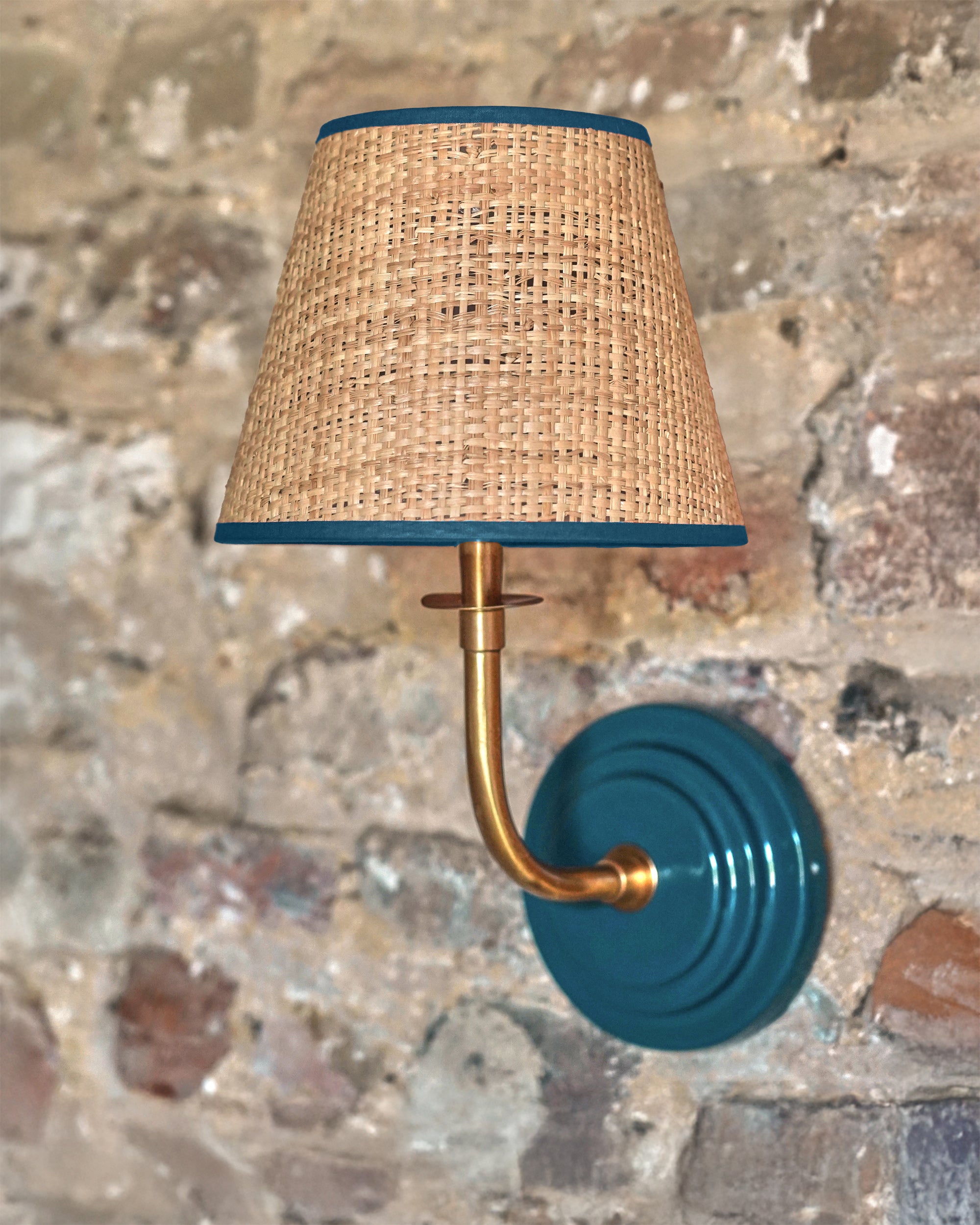 Wall-mounted lamp with wicker shade and teal base on a textured stone wall.