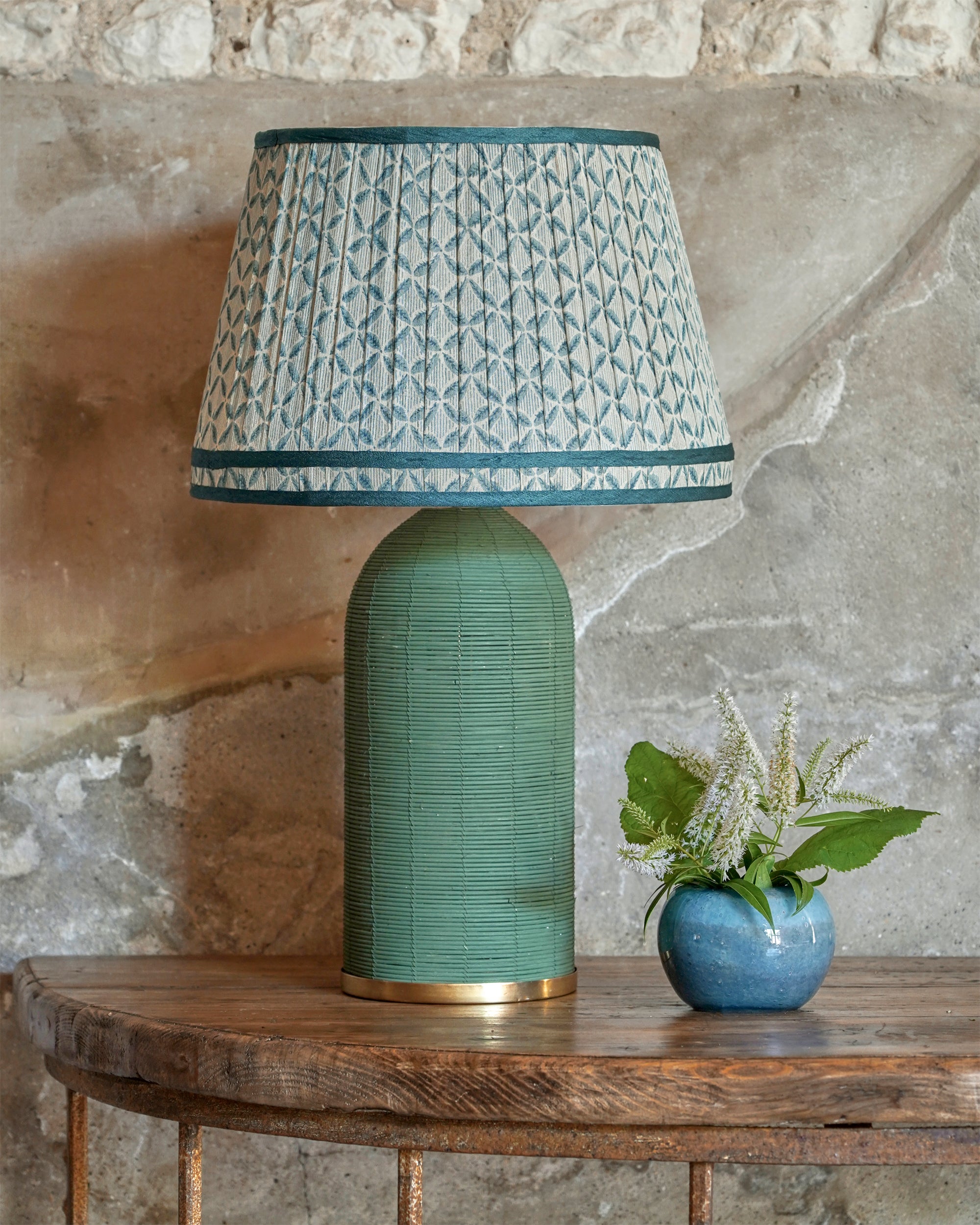 Green textured table lamp with patterned shade on a wooden table against a stone wall.