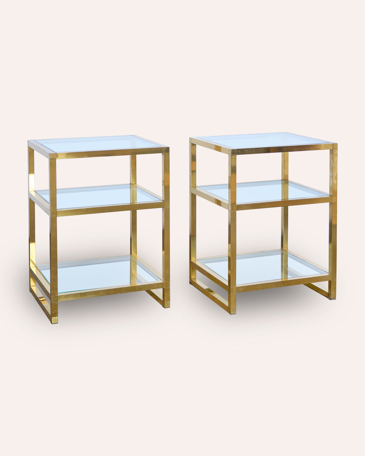 A pair of modern three-tier side tables with brushed gold metal frames and light blue glass shelves, shown against a solid pale beige background.
