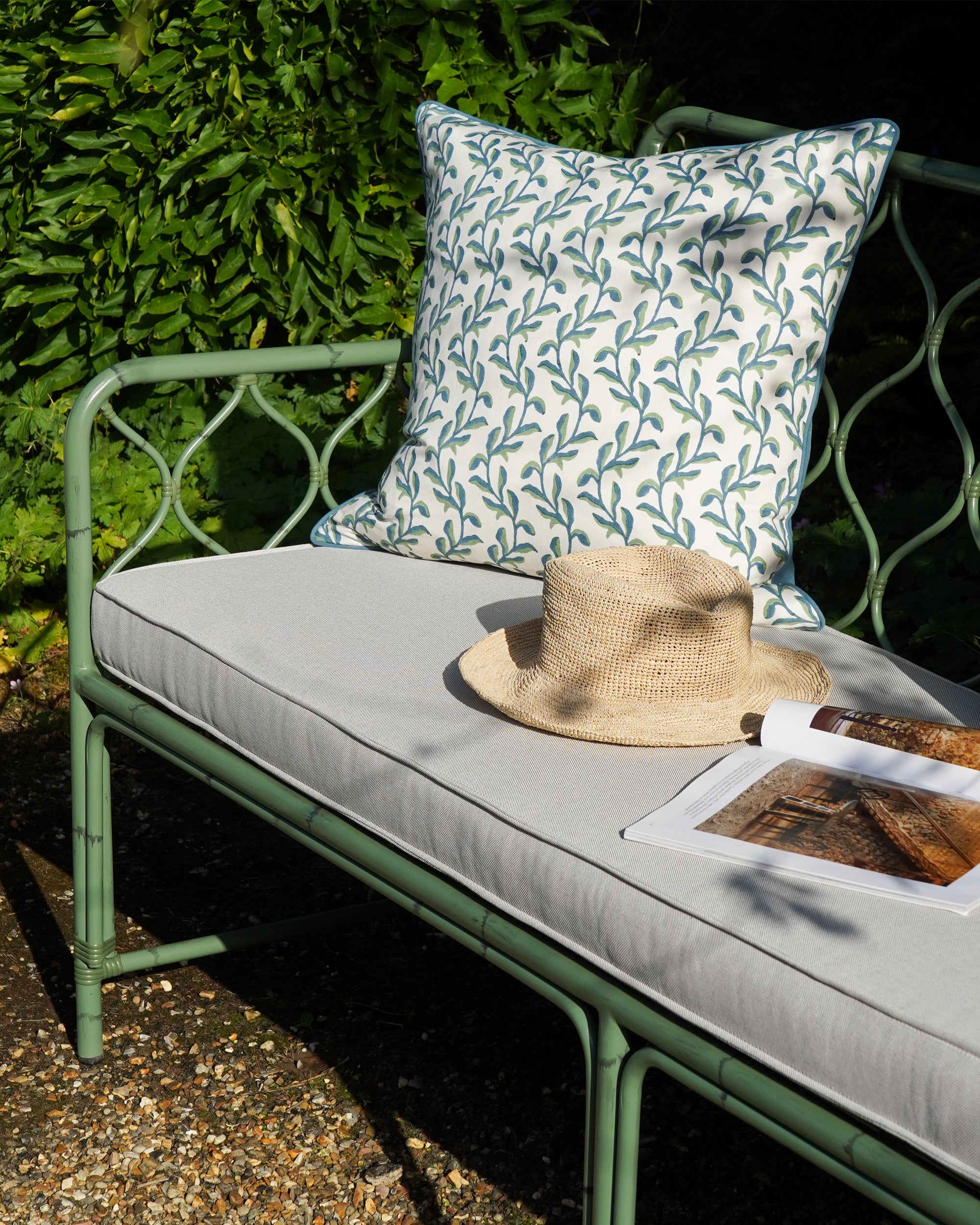 Green Curato sofa with floral blue and green patterned cushion, straw hat, and magazine in a garden setting.